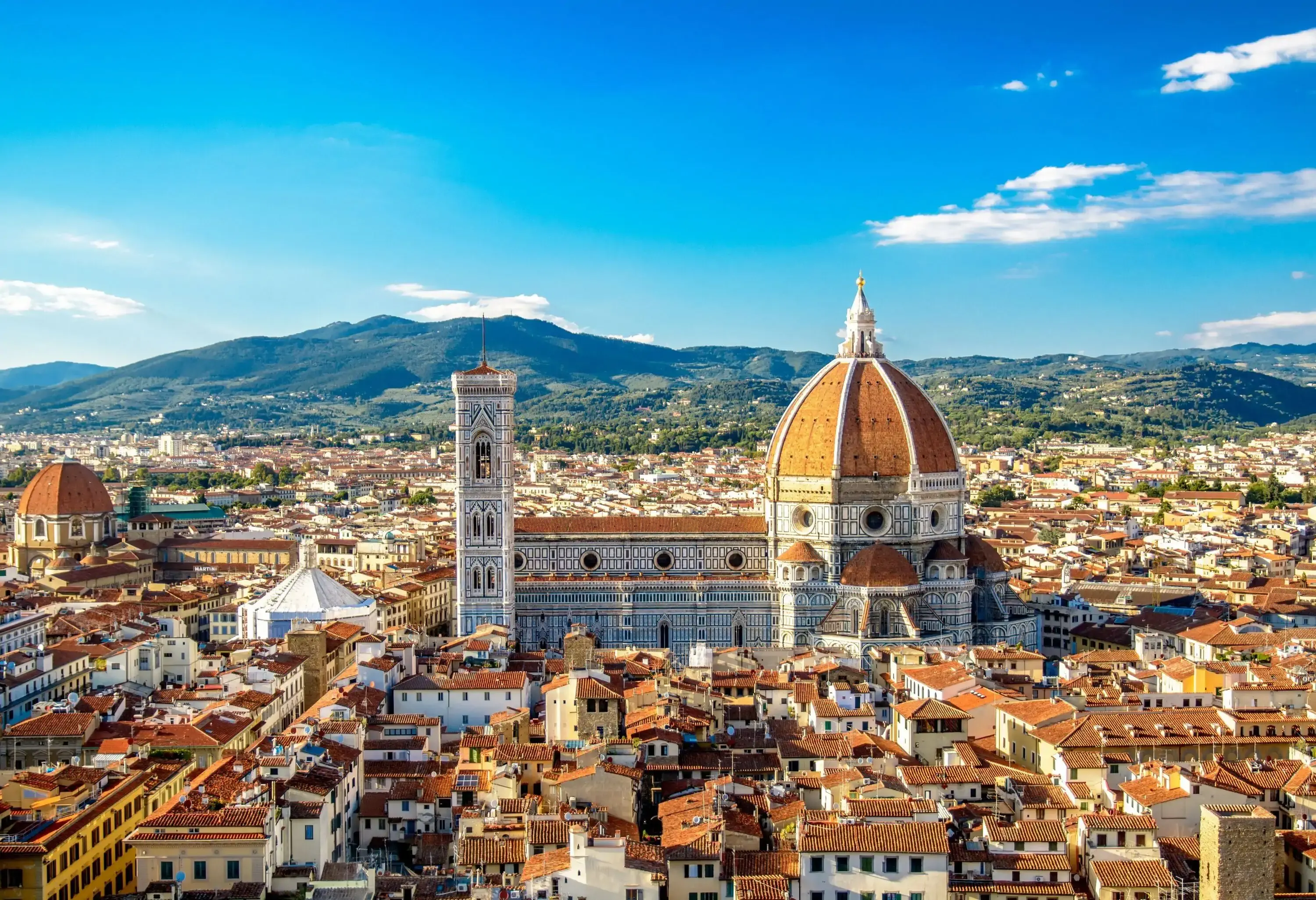 A stunning gothic-style cathedral with a red-tiled dome in the centre of an old town surrounded by traditional buildings against the lush mountain range.