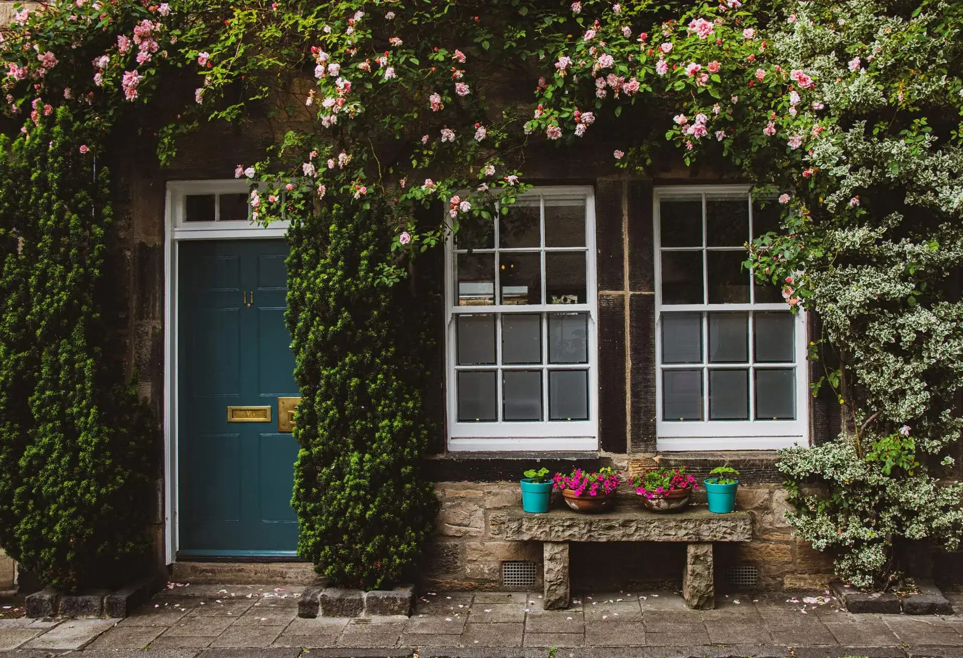 The front of a stone house is covered with flowering vines and flower-filled pots atop a stone bench.