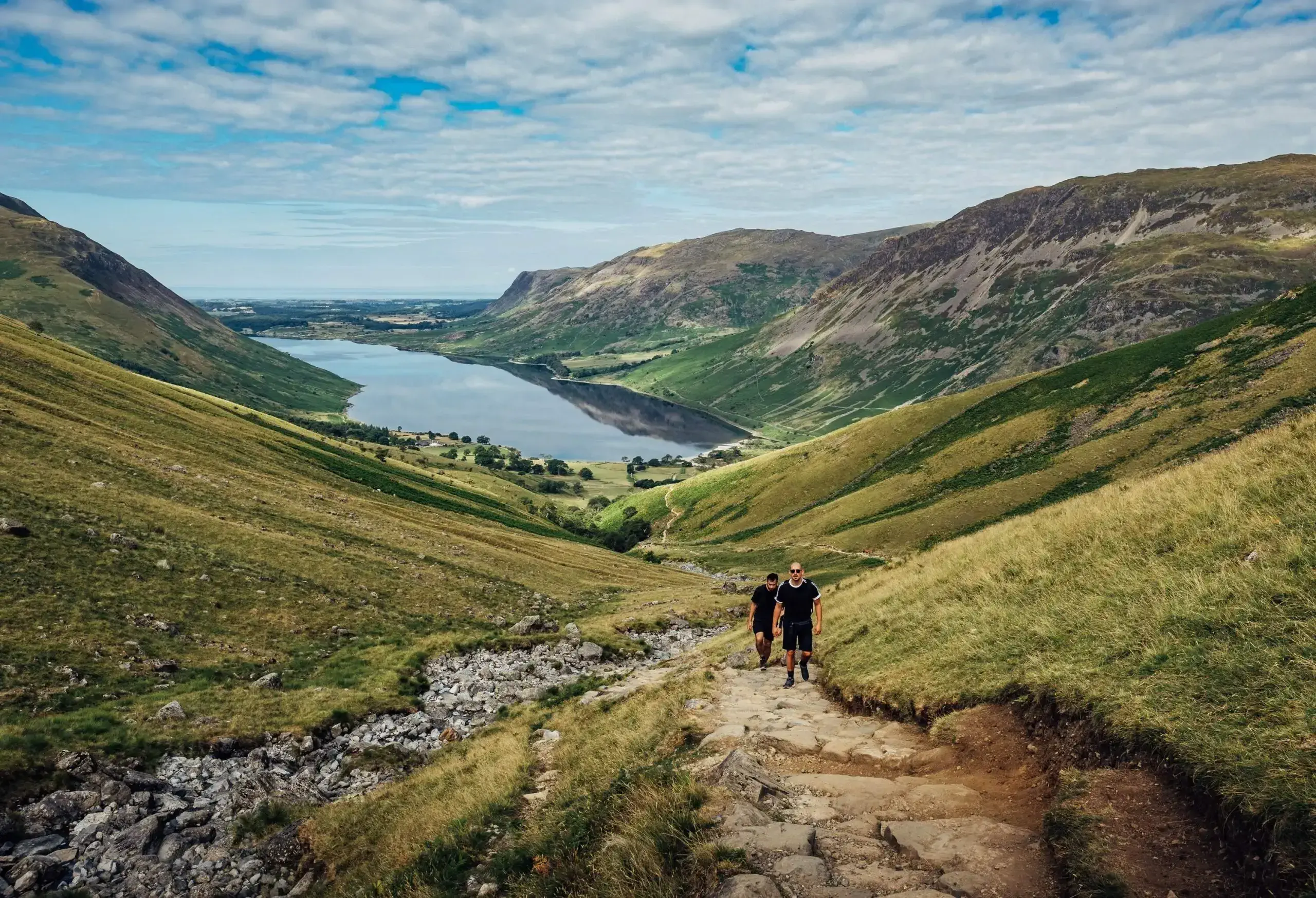 Two individuals ascending a stone staircase between two grass valleys with a lake in the distance.