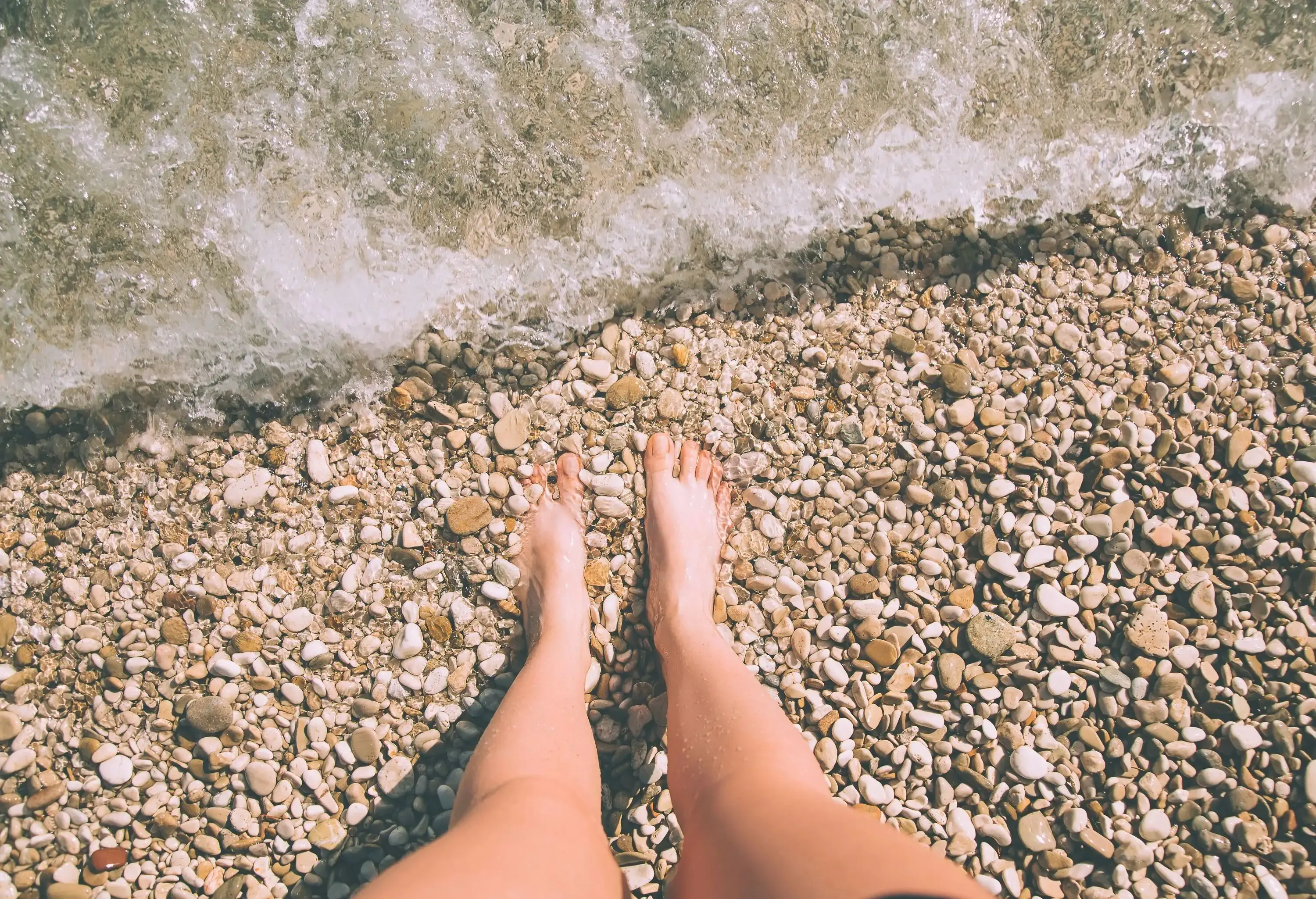 A woman's feet firmly planted on a pebbly beach splashed by the waves.
