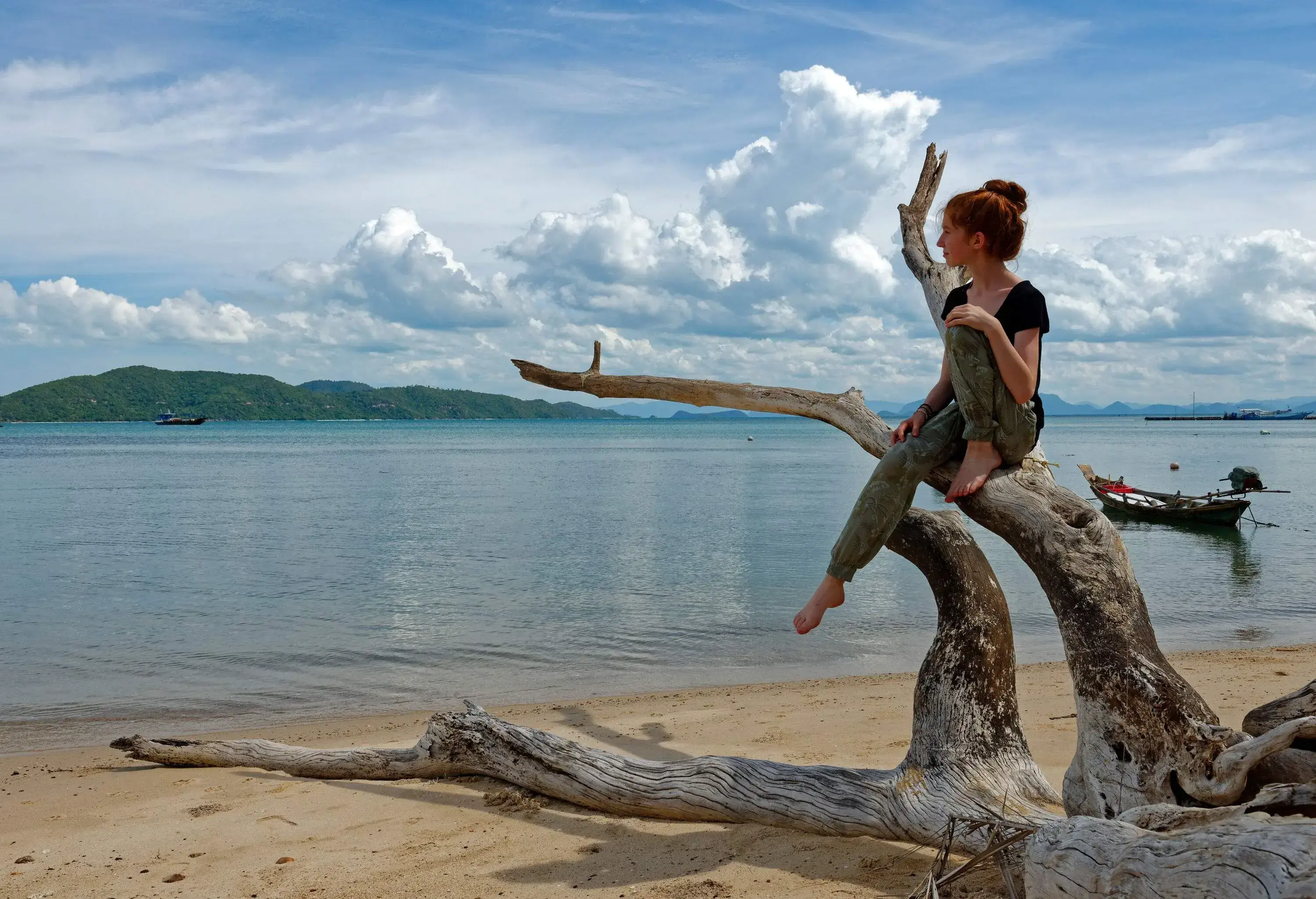 A woman sits on a dead tree trunk on the shore of the beach.