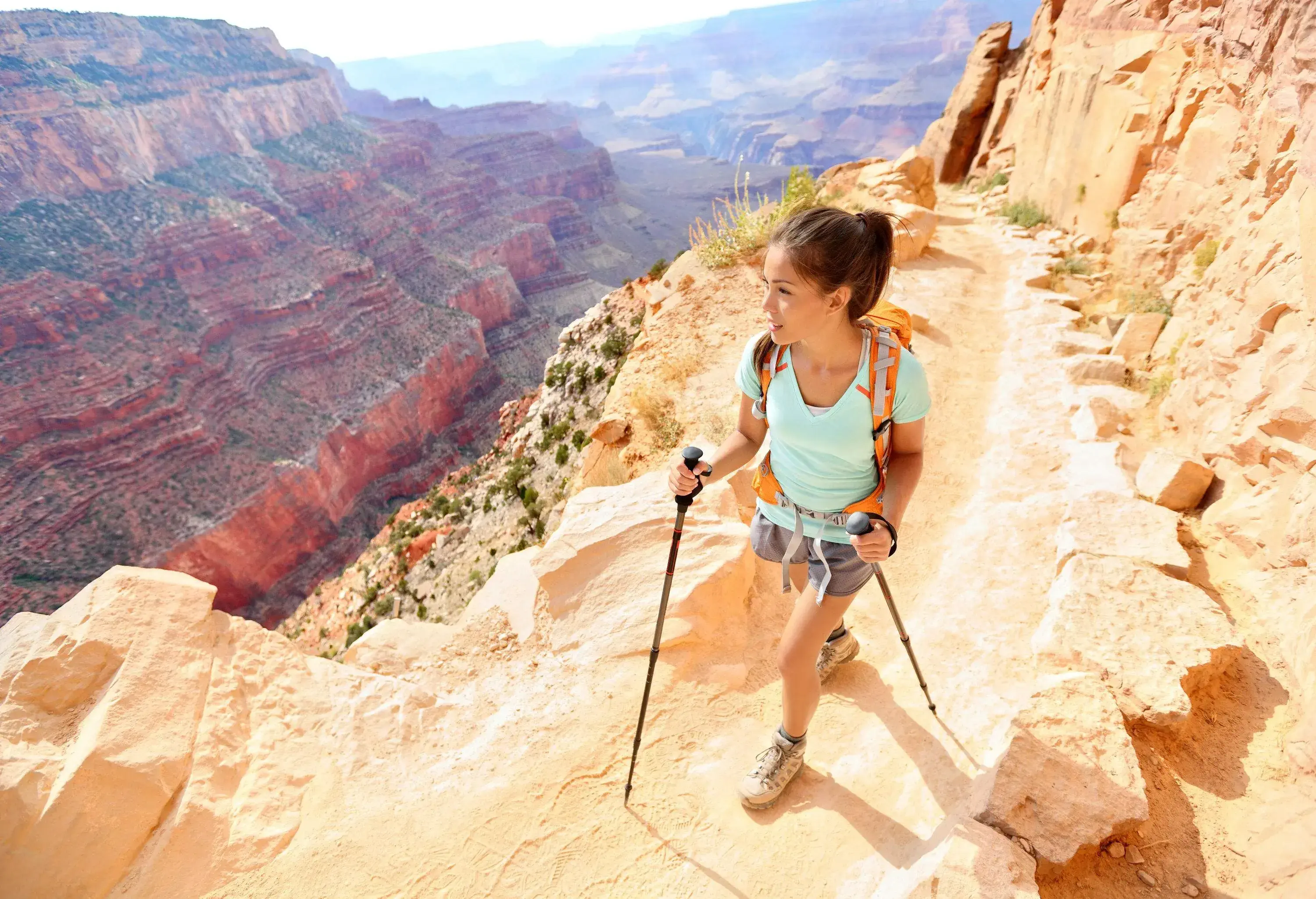 A woman clutching trekking poles walking on a narrow path carved into the rocky mountains of the Grand Canyon, Arizona, USA.