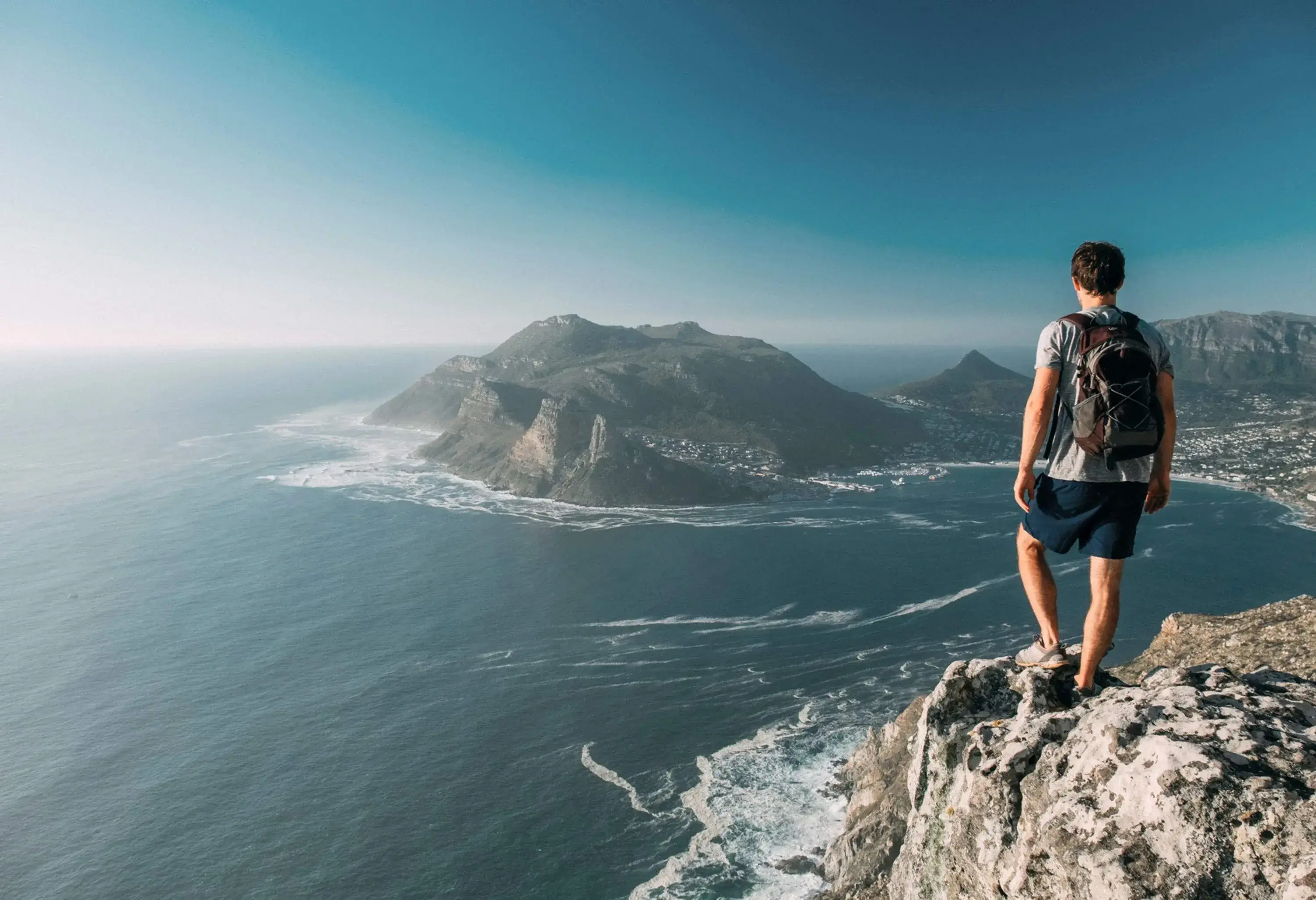 A man with a backpack stands on a rock cliff and stares at the vast ocean, coastal city, and surrounding mountains below.