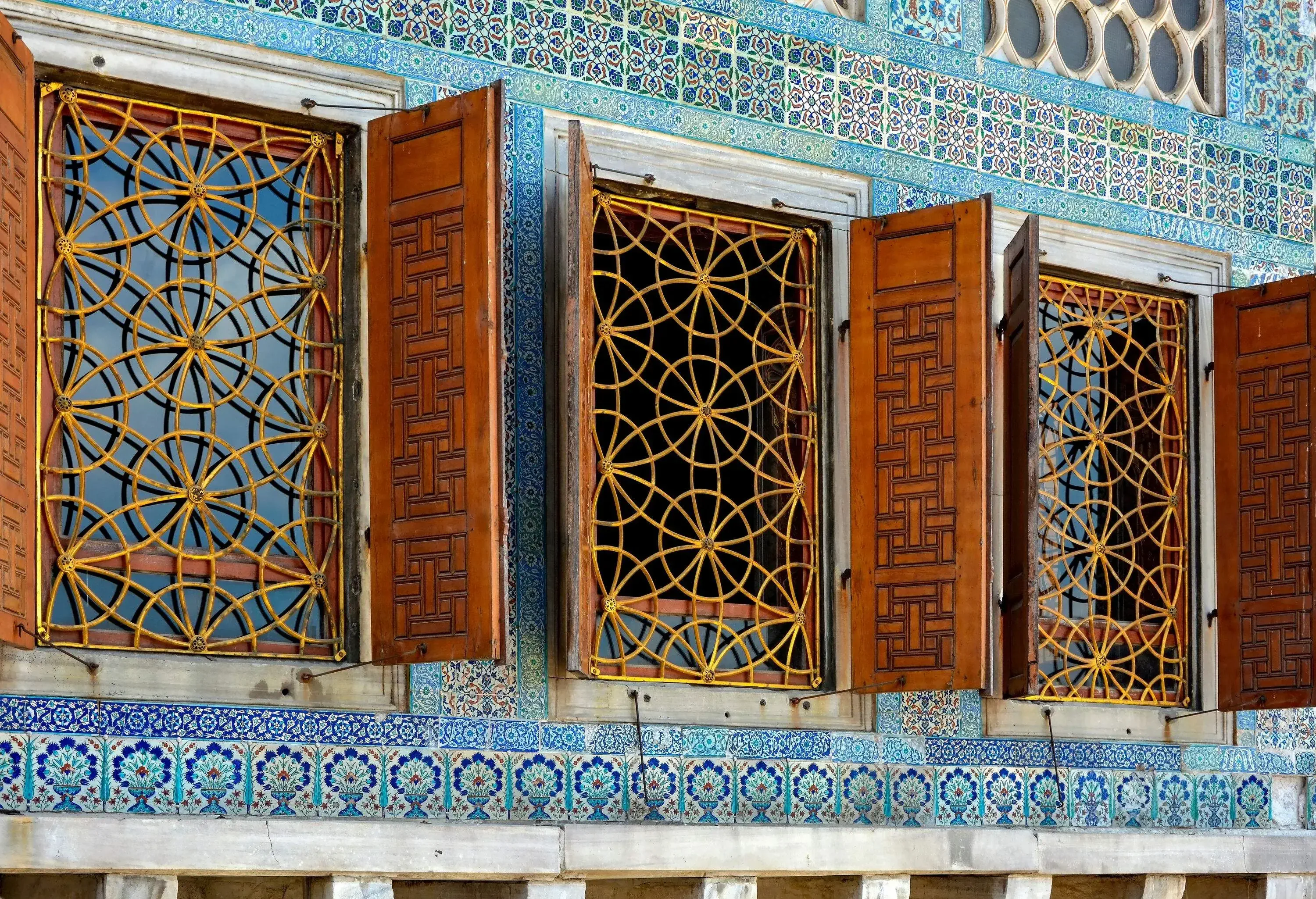 Three wooden glass windows with golden grills in a blue-tiled wall.