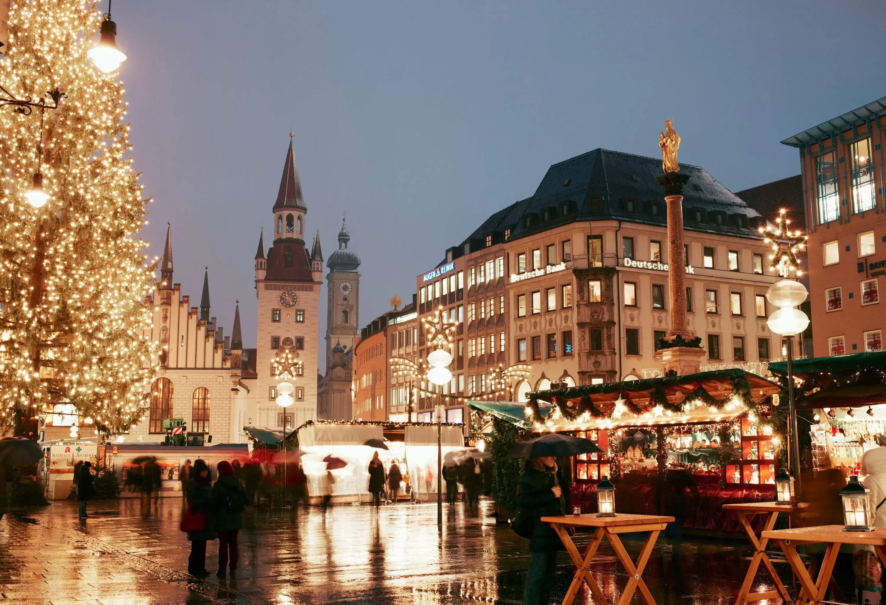 A giant illuminated Christmas tree and Christmas market in the central square of Munich, Germany with a view of the Old Town Hall on the back, on a cold rainy evening.