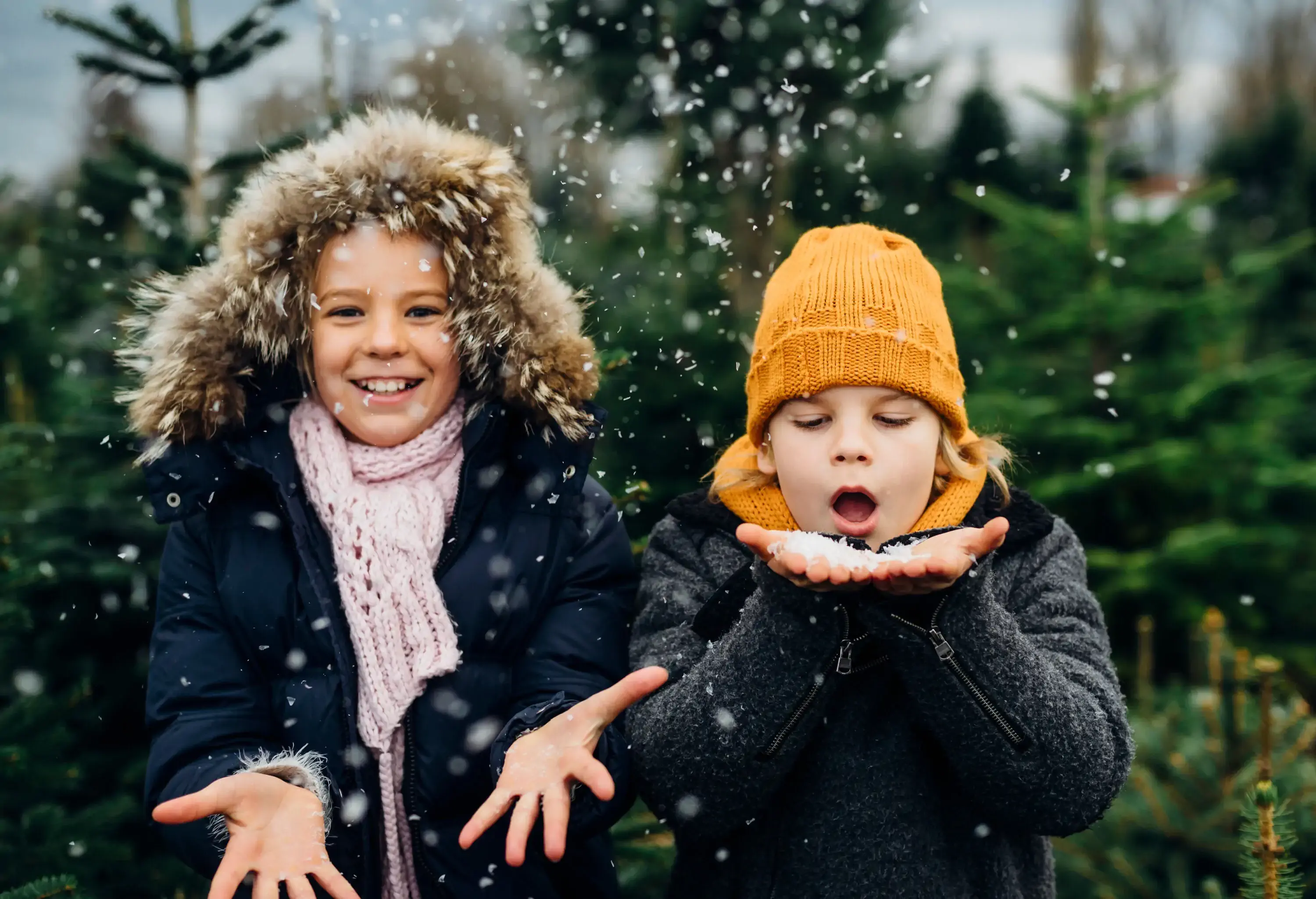 A happy child in a parka held out his hands to catch the snow being blown from the hands of a child wearing a yellow knitted cap.
