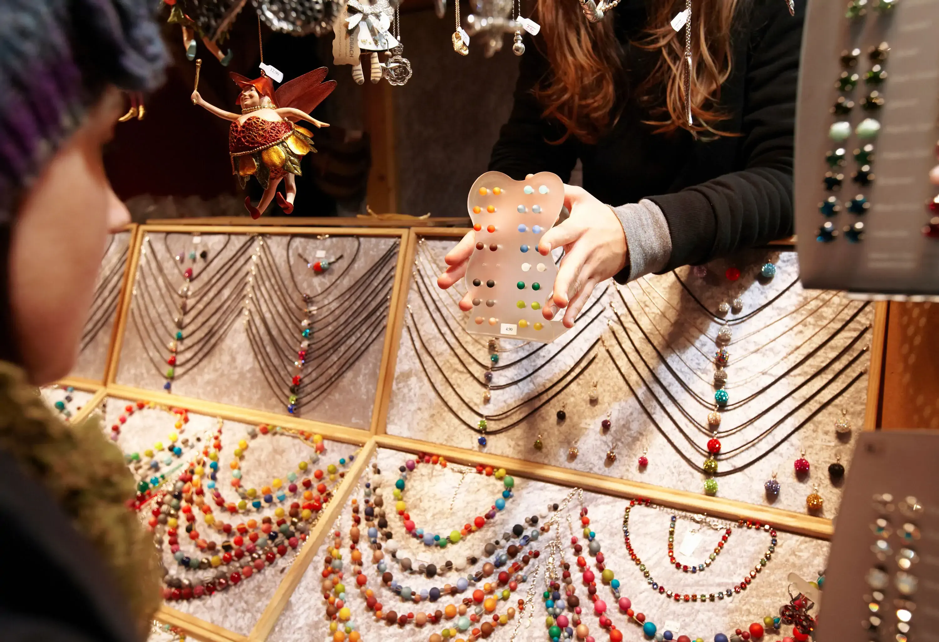 Hands holding a selection of jewellery earrings in a shop with a display of colourful necklaces.
