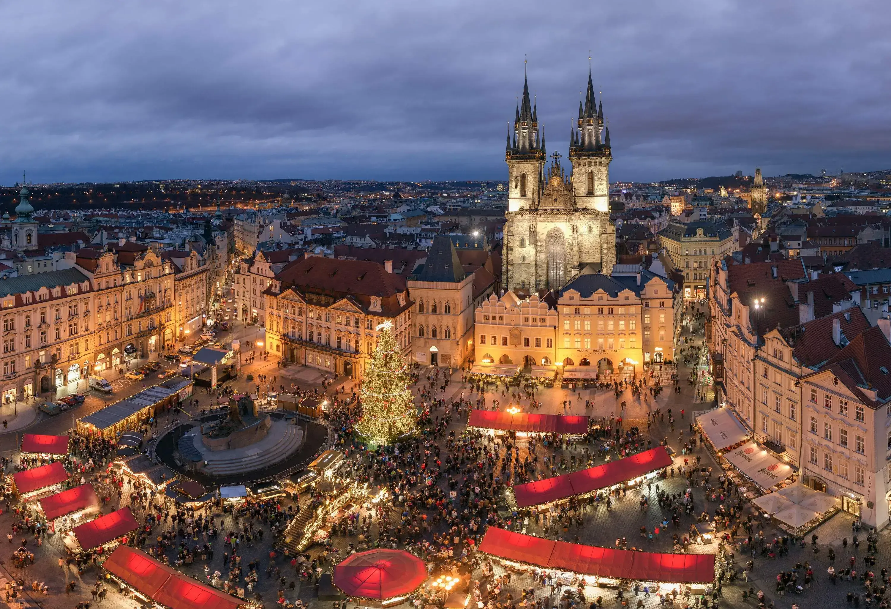 Night-time scene of a town square's Christmas market surrounded by buildings and a Gothic church with towers of multiple turrets.