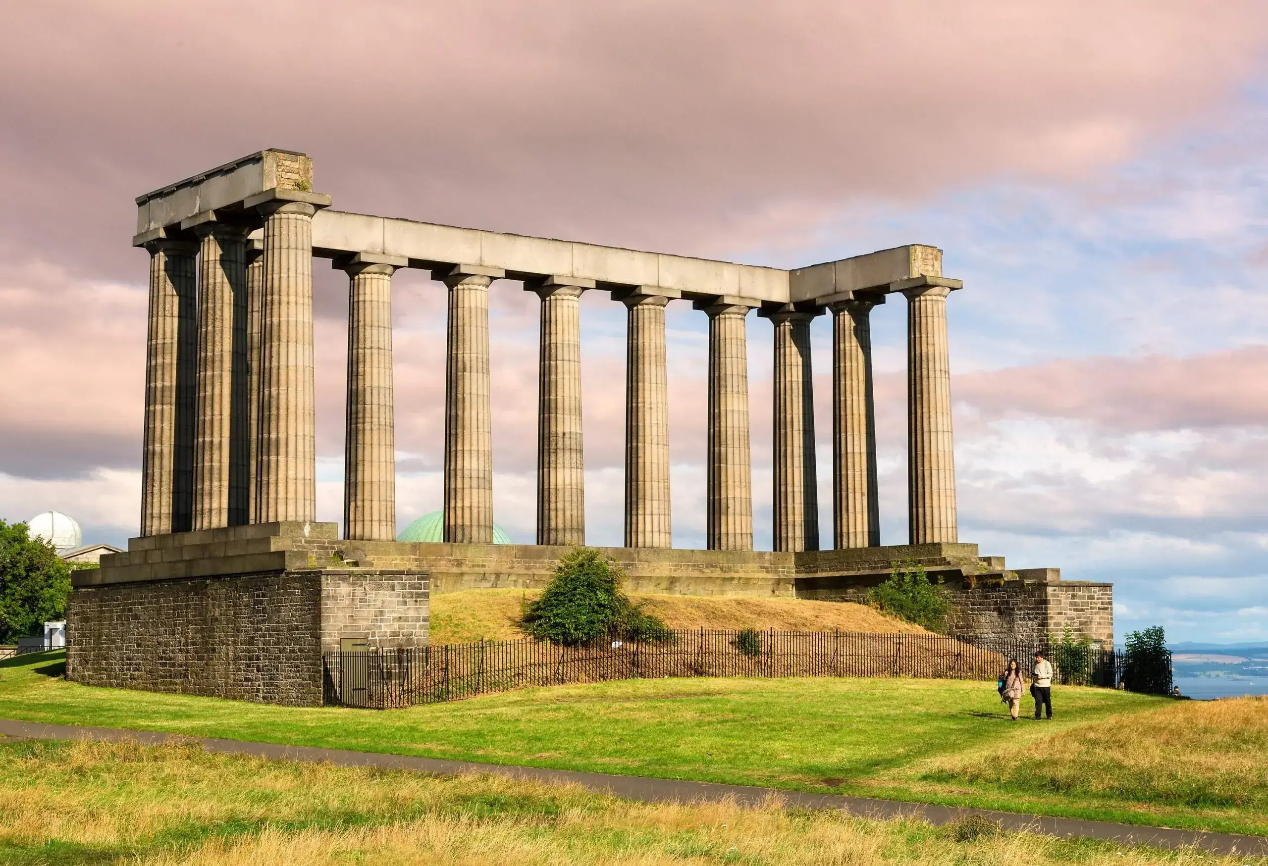 Two people stroll across a grass field next to a pillared monument.