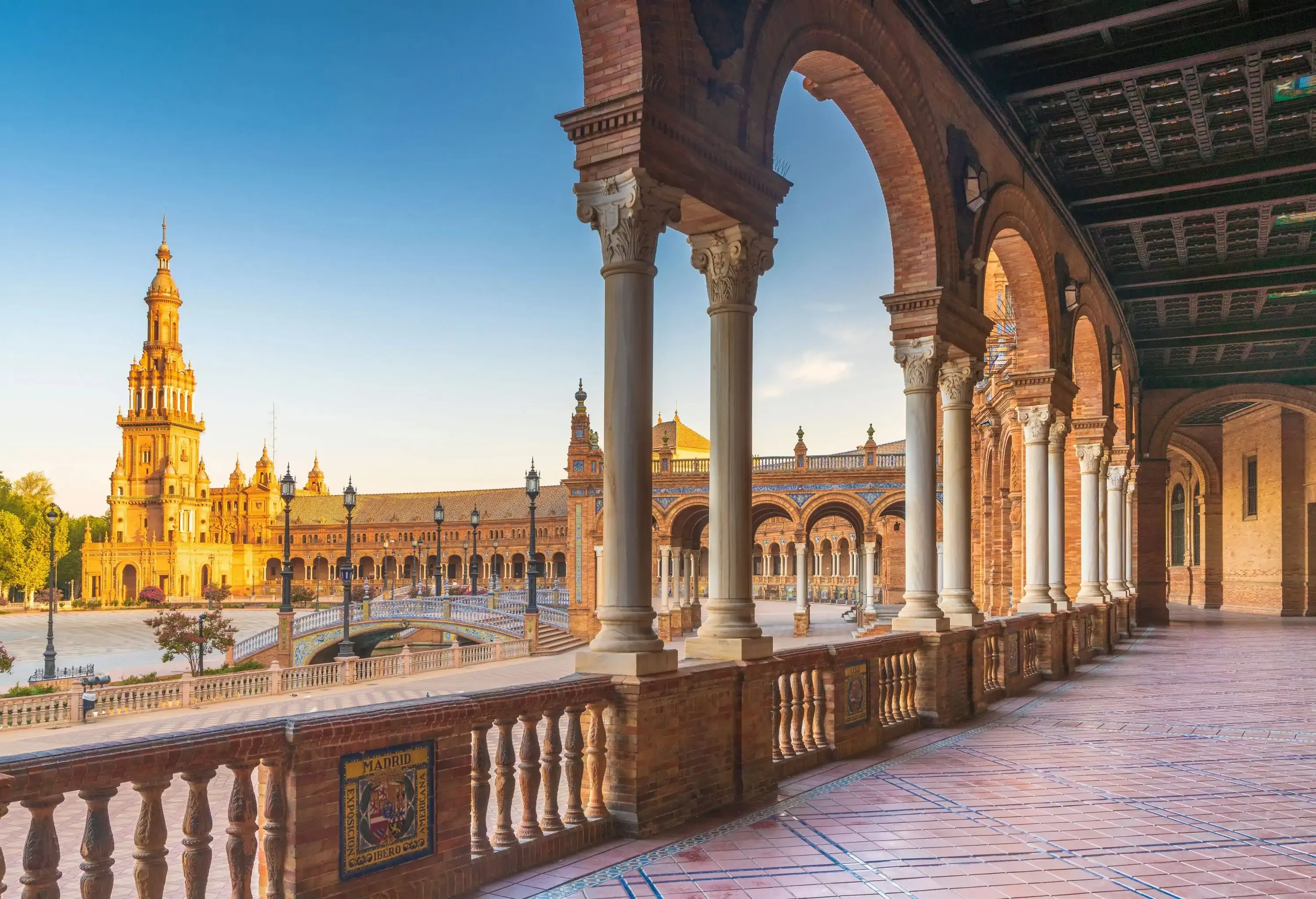 An antique tower at the end of a long-span building is visible from the colonnade of Plaza de España's round portico.