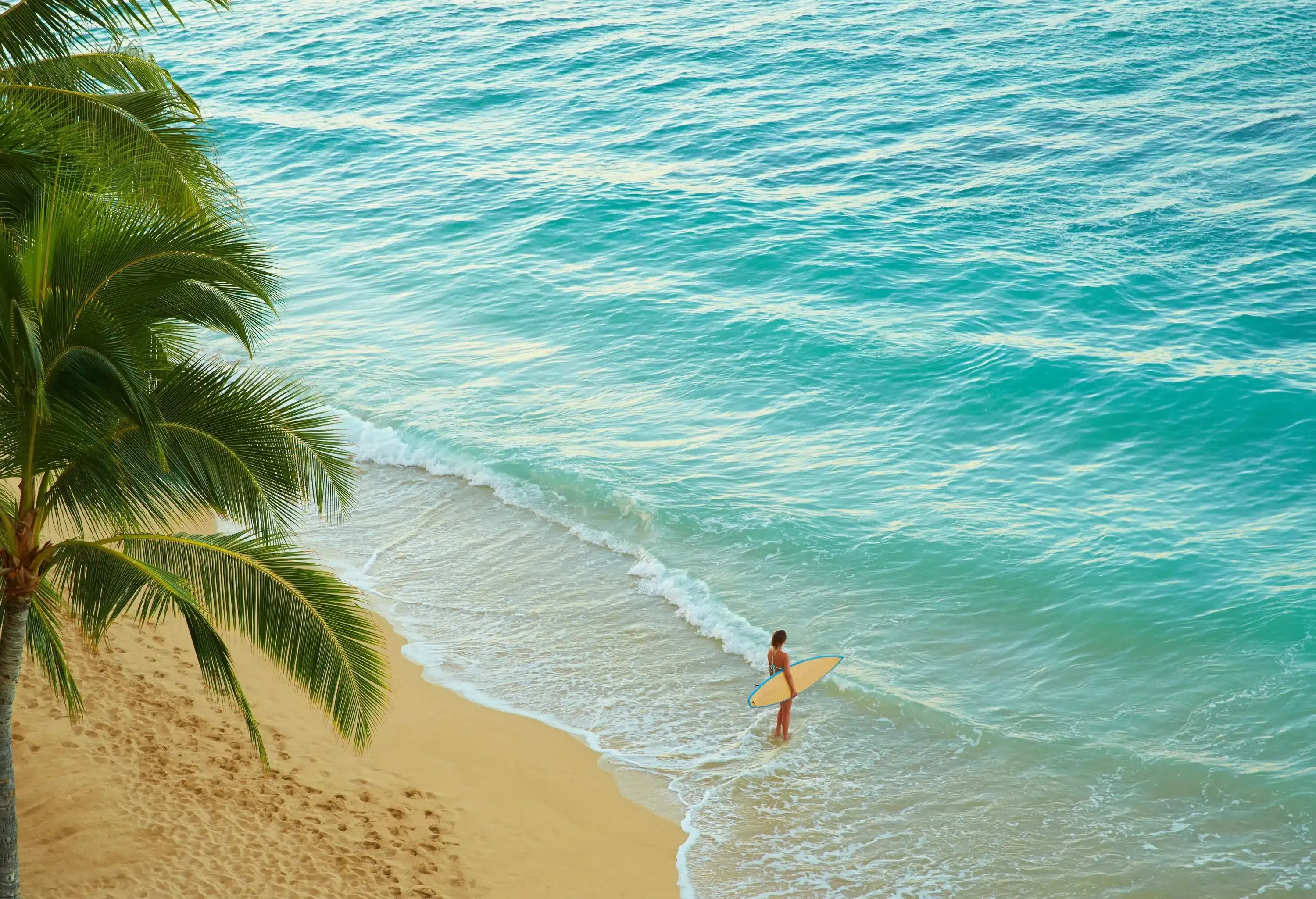 A woman standing on the beach with a surfboard while the waves smash on her feet.
