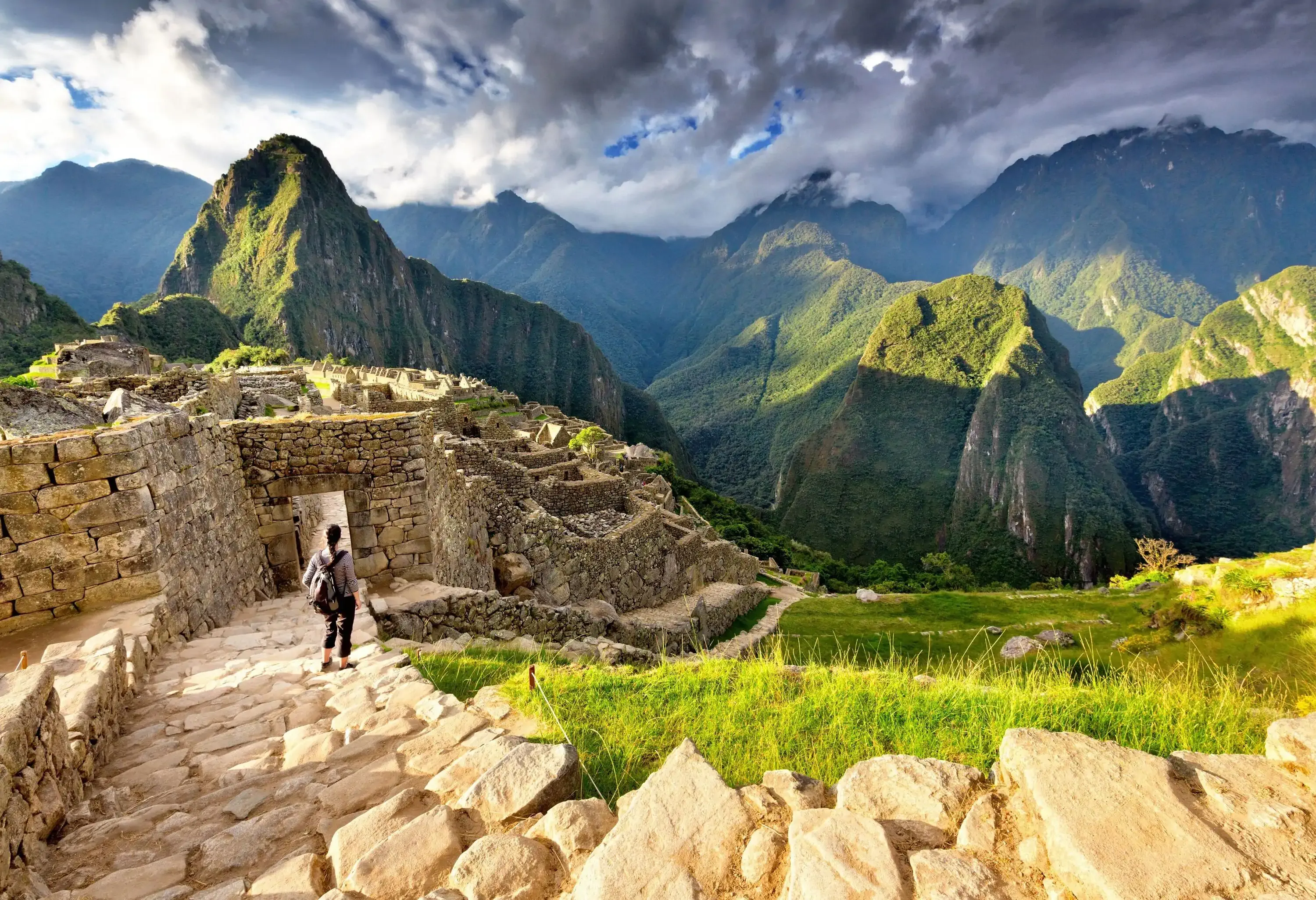 A stone-paved walkway gracefully descends towards ancient ruins, as a person carrying a backpack makes their way down, overlooking a breathtaking vista of a verdant mountain range.