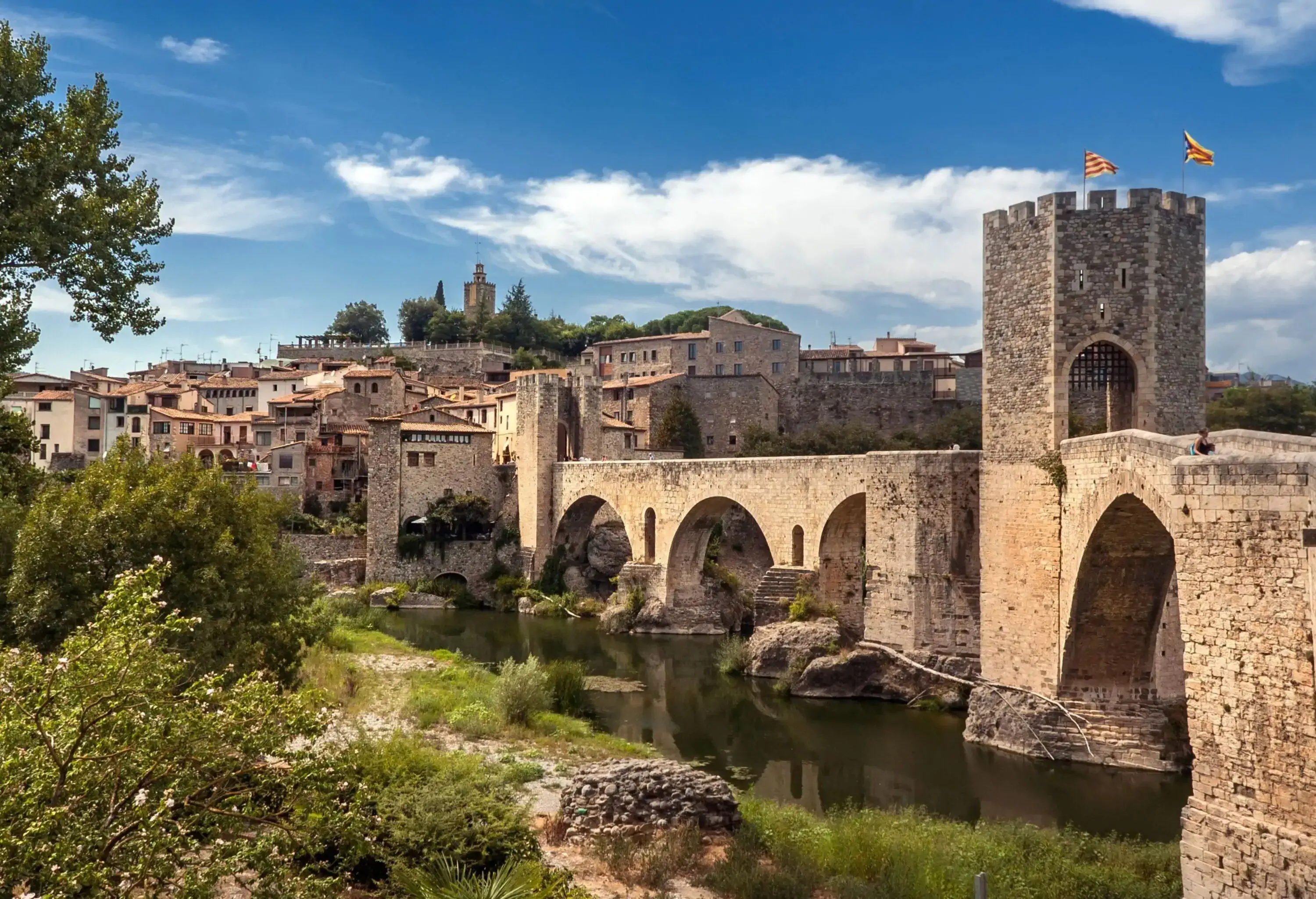 An old arched bridge mounted on rocks connected to a village on a hill through a tower.