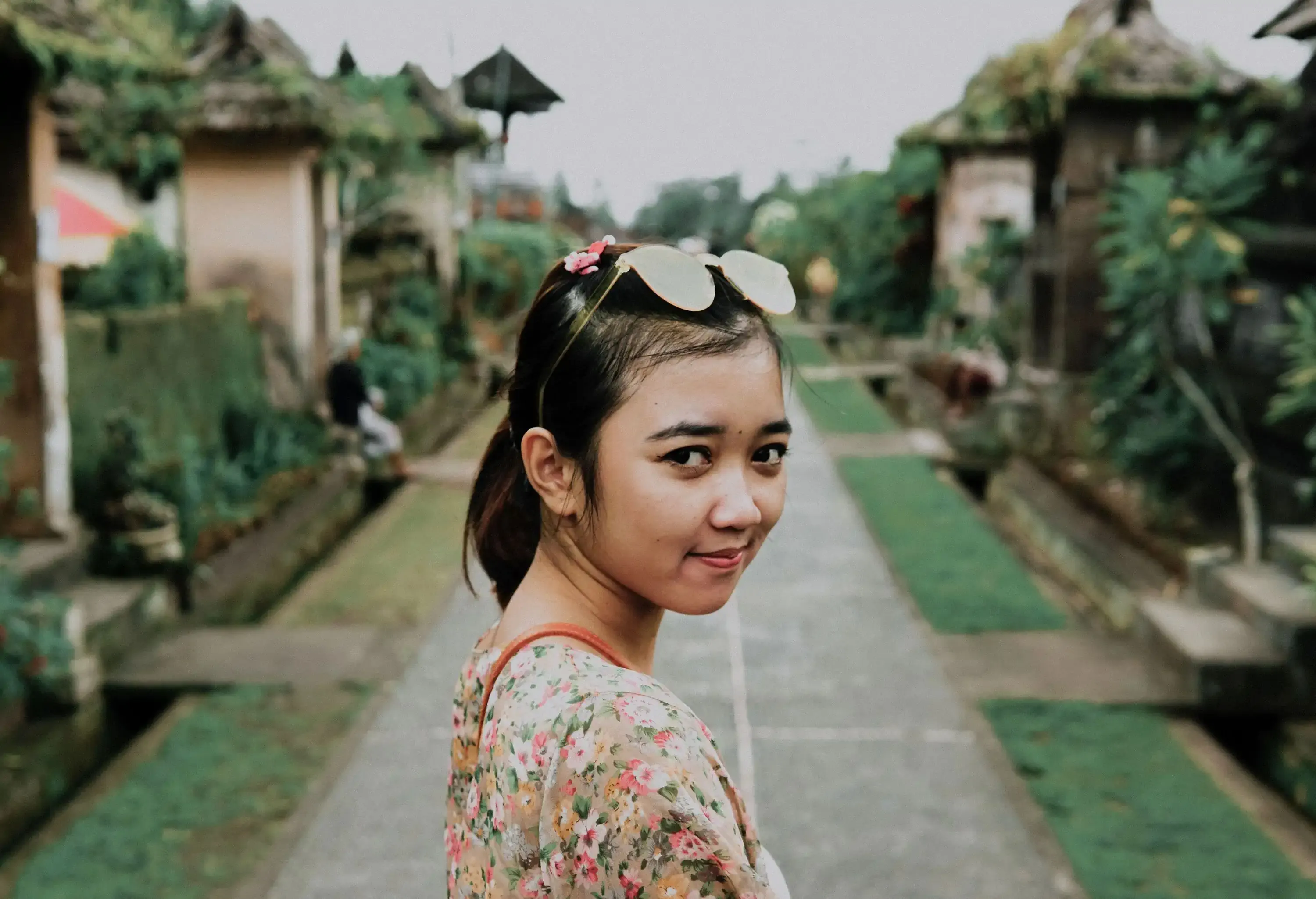 A woman in a floral t-shirt with sunglass rests on her head against a blurred lush background.