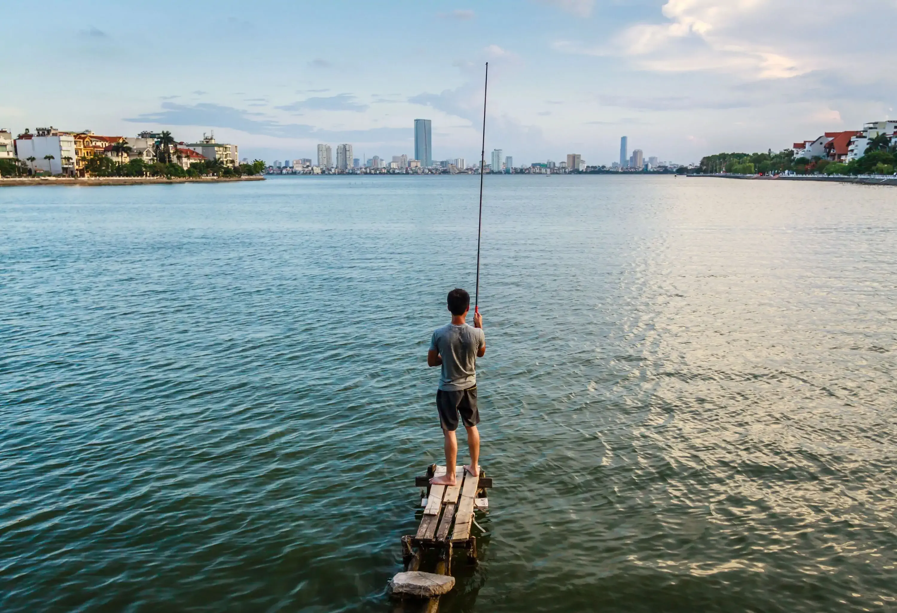 A man stands on a wooden pier in a lake while holding a fishing rod.