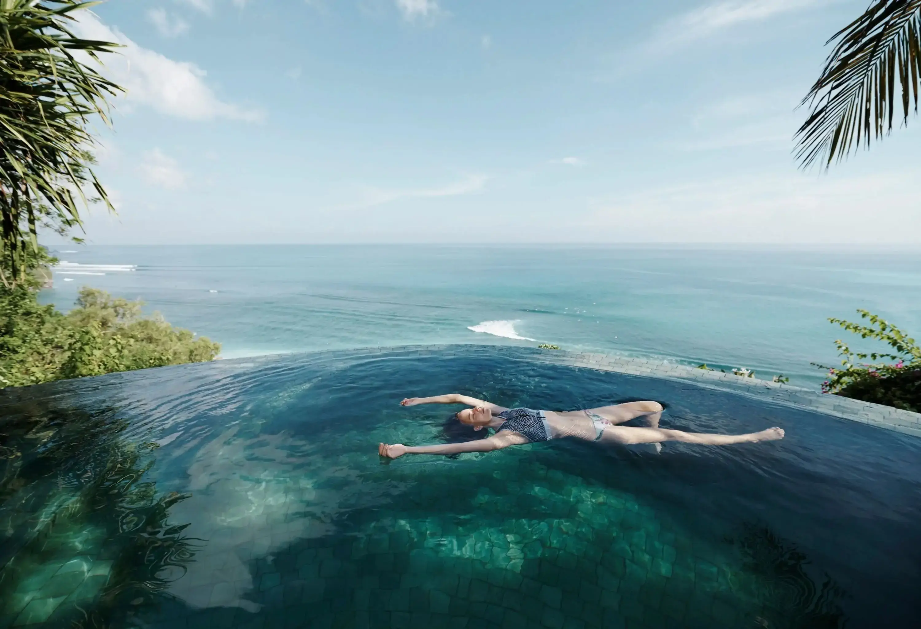 A woman floating on her back in an infinity pool facing an expansive ocean.
