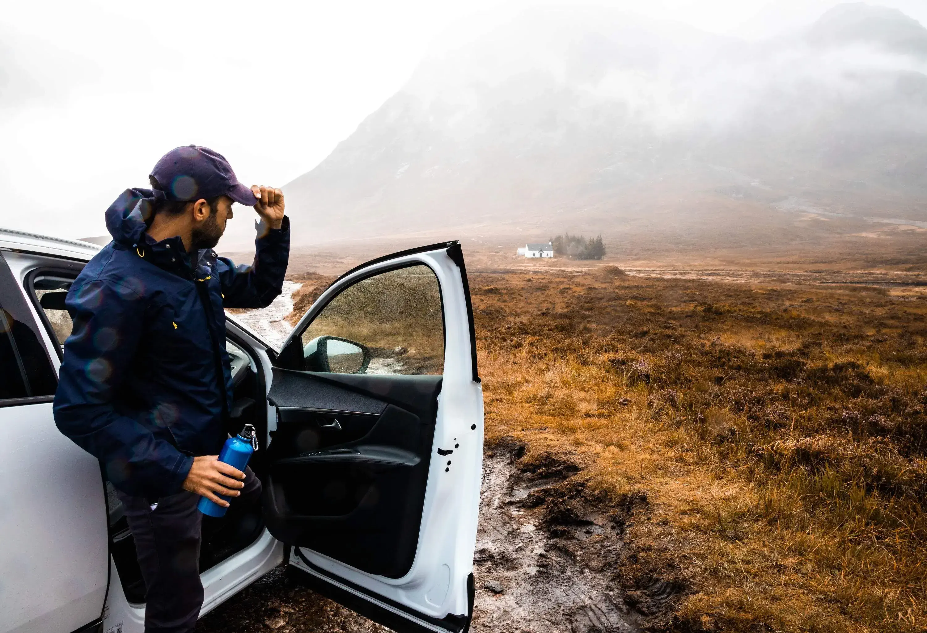 Man clad in a blue windbreaker emerges from a white car, holding a flask and looking at rugged landscape on a cloudy day