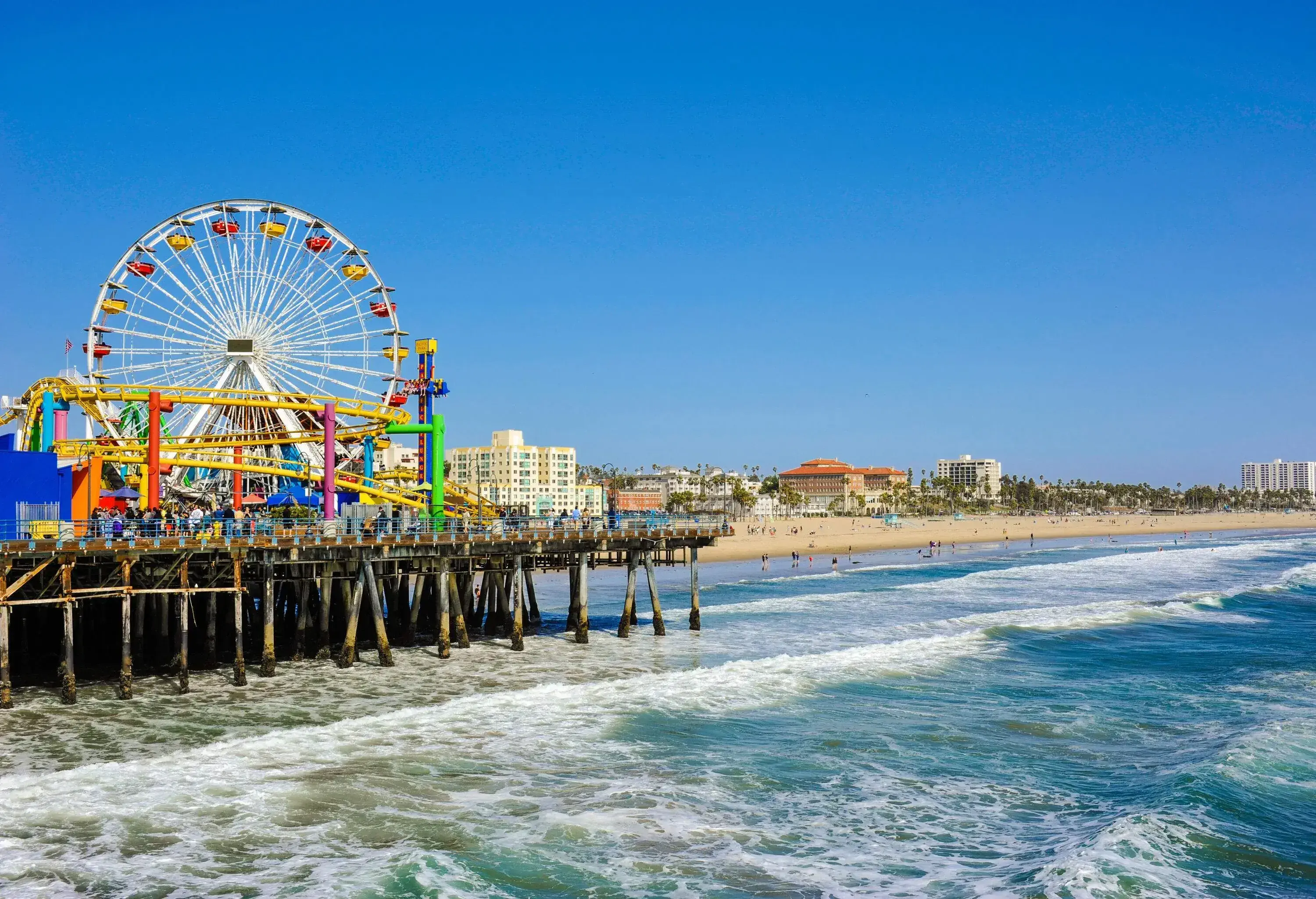 A pier with a colourful amusement park on the shore of a beach along an urban cityscape.