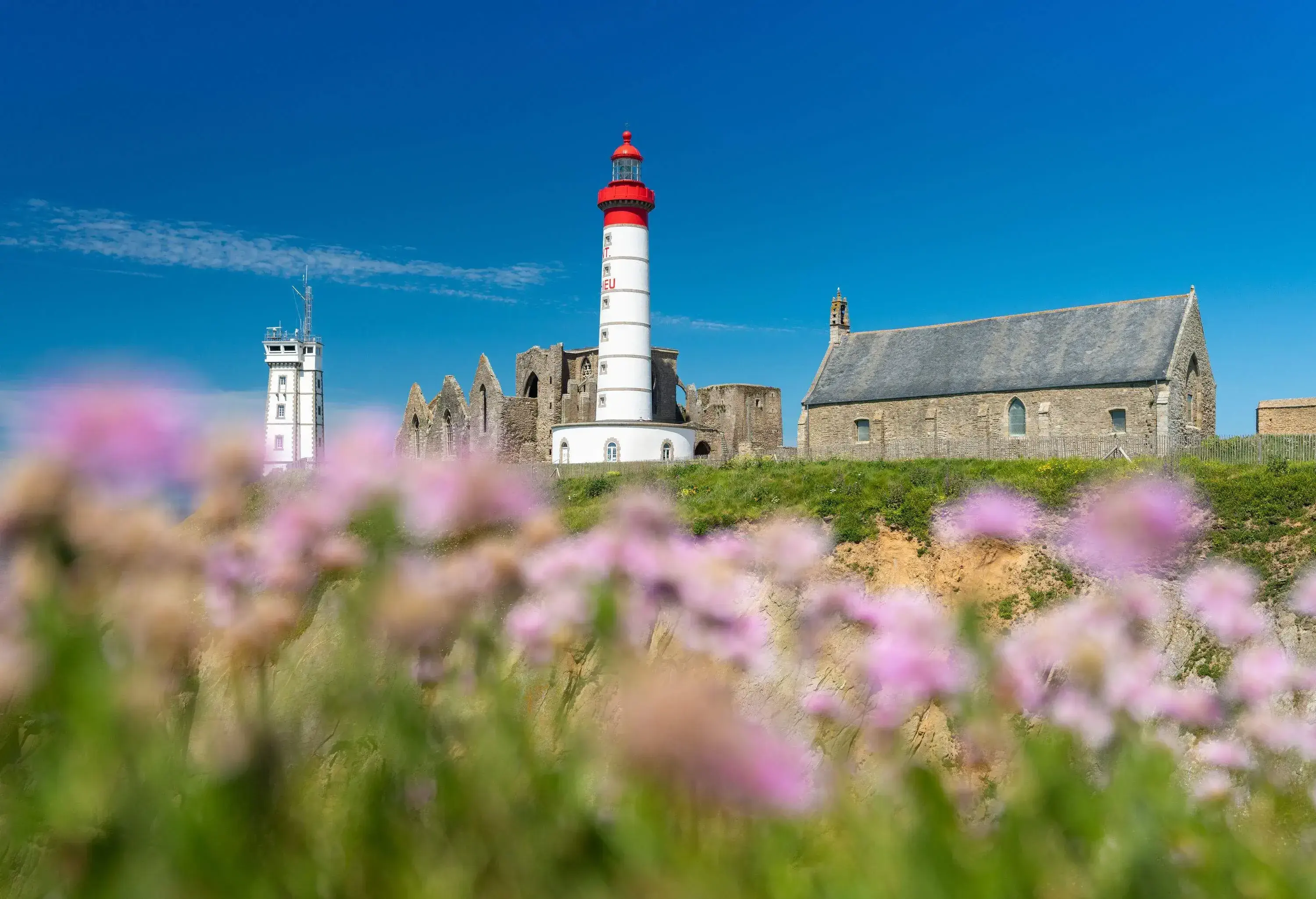 A lighthouse beside the ruins of an abbey and a signal station in the background over a lush green land.