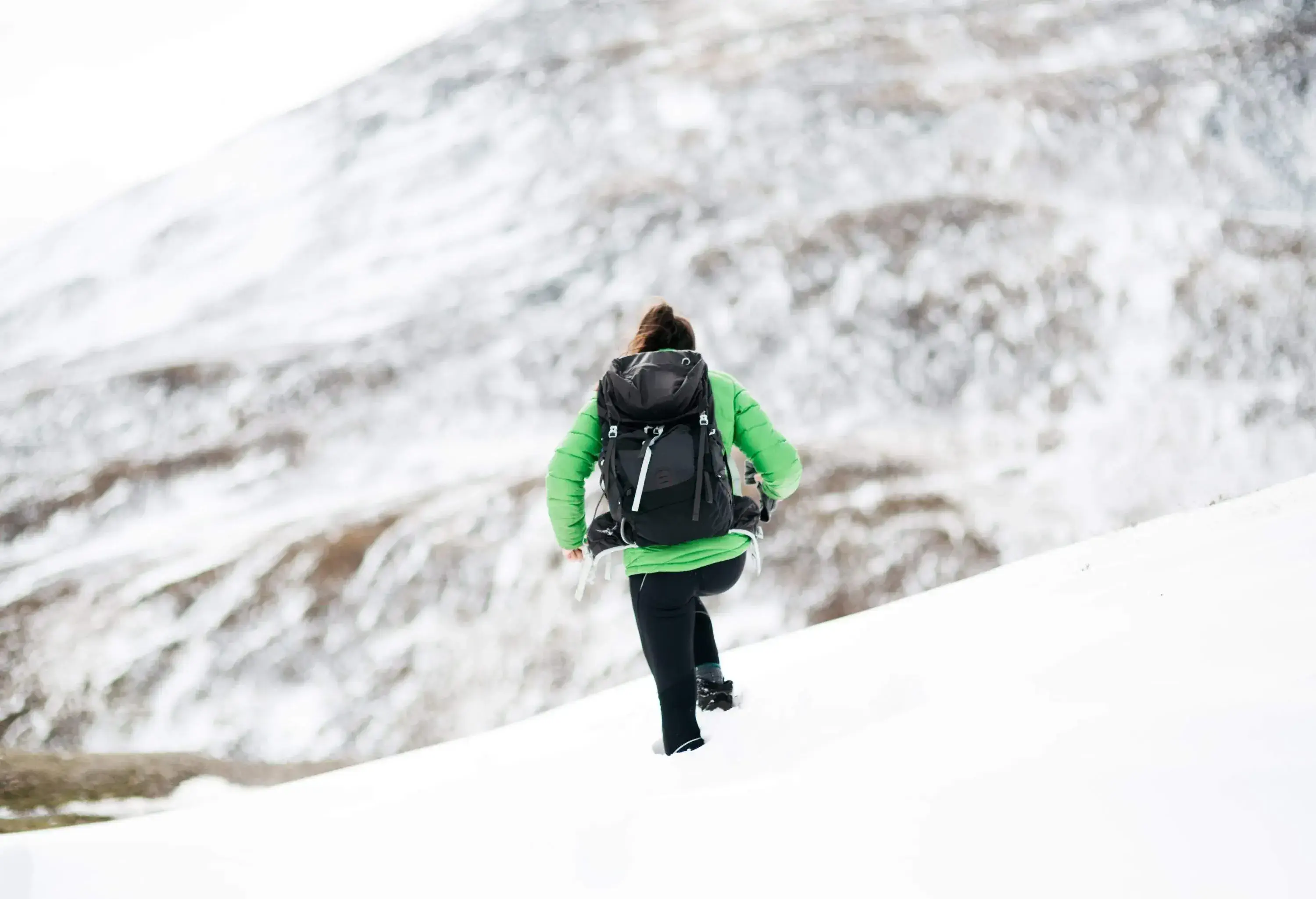 Woman wearing green winter jacket and backpack hiking in the snow by the mountains