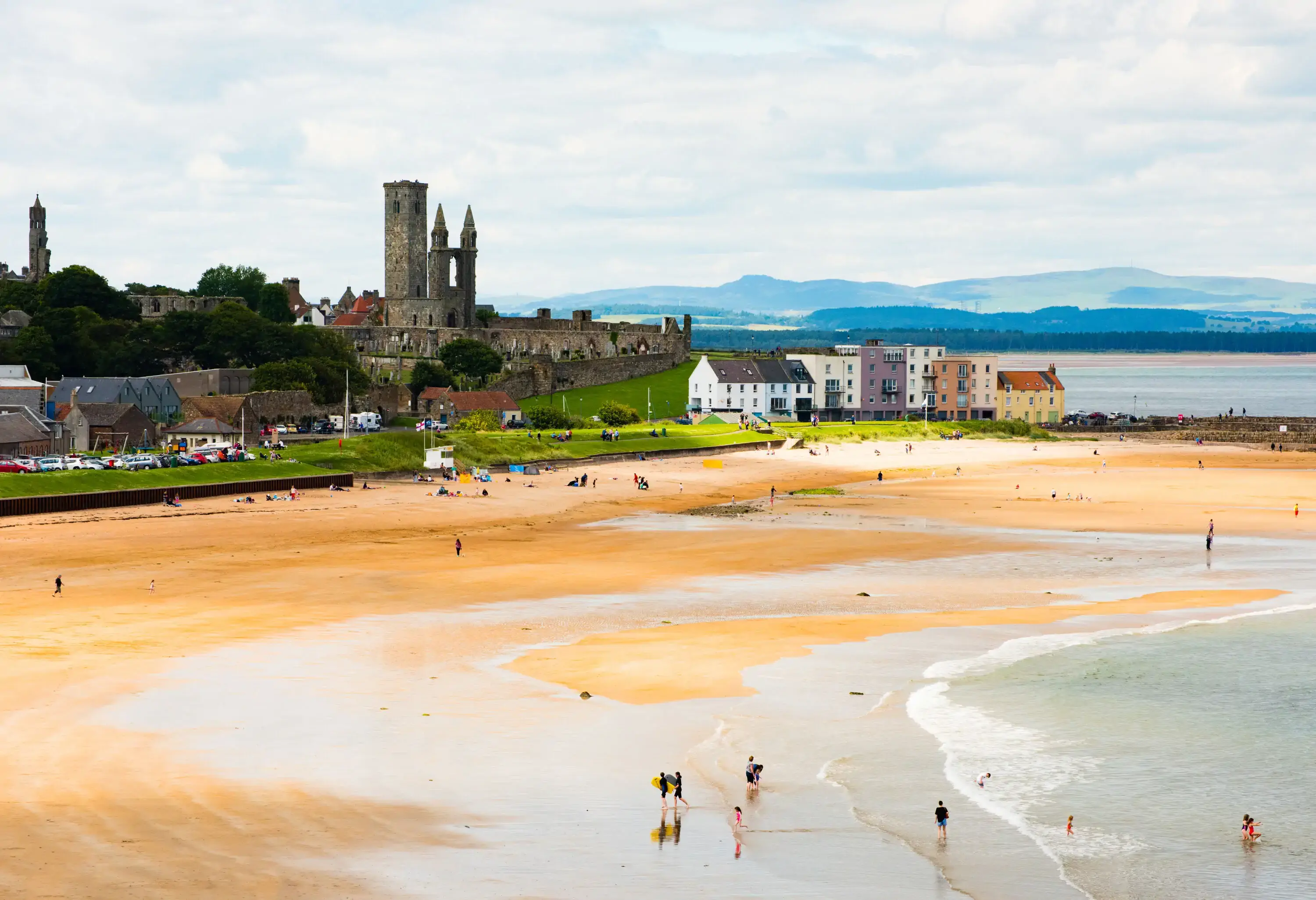 People walk across a beach along waterfront buildings with a distant view of a ruined church's square tower and twin steeples.