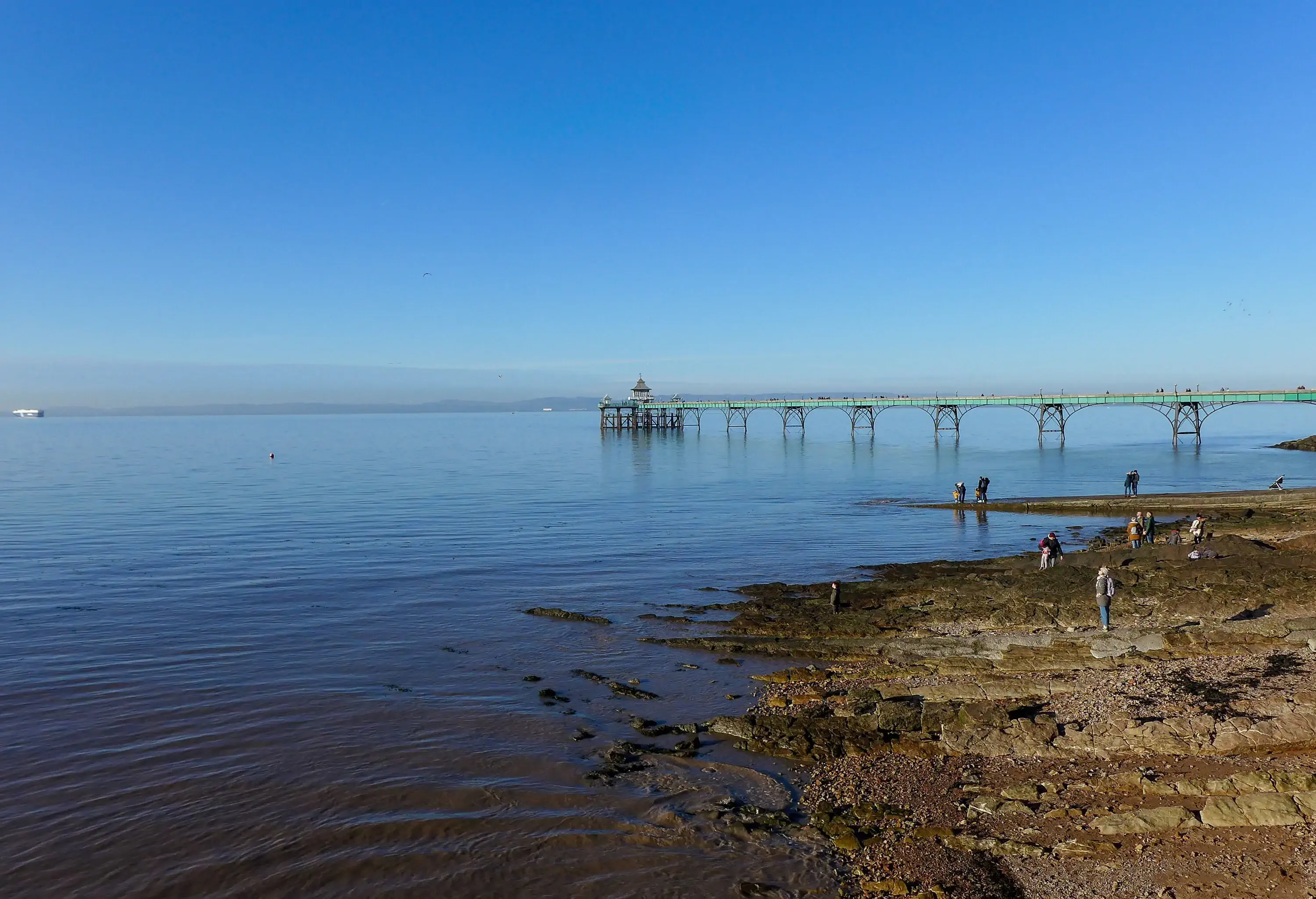 People on the rocky shore overlooking a historic Victorian pier over the tranquil sea.