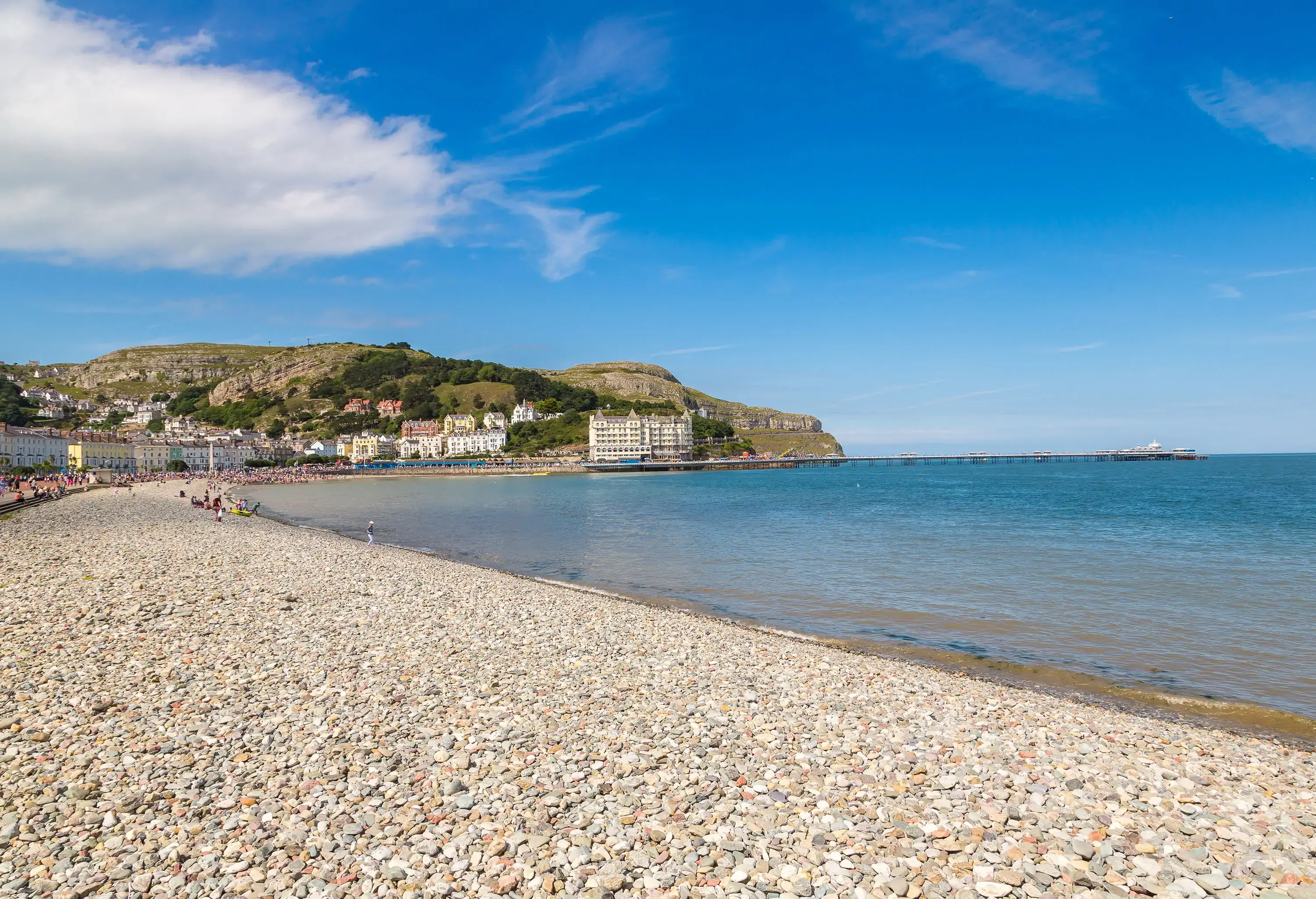 A shingle beach shore lined with tall and modern buildings beneath a lush hill.