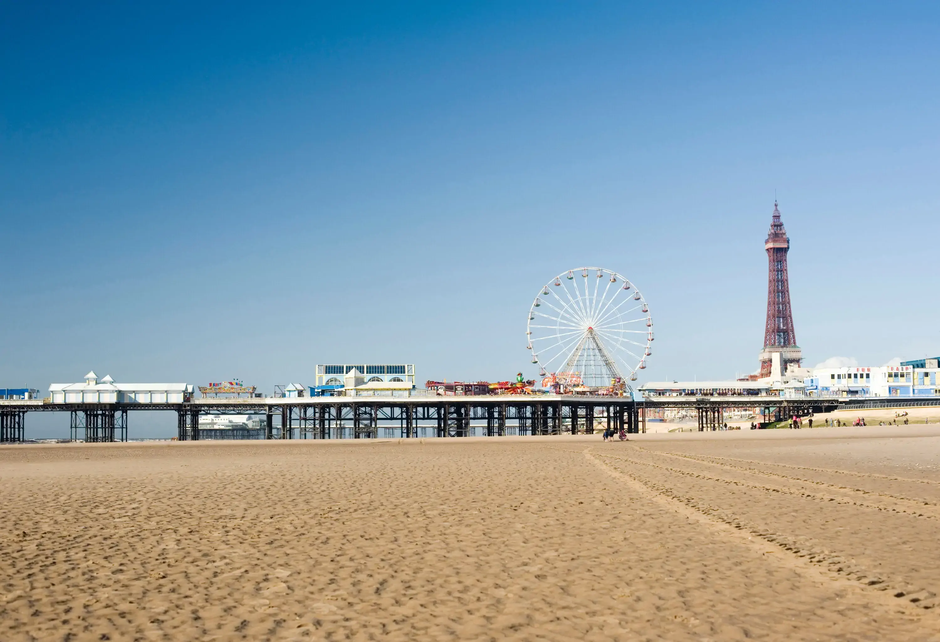 A beach with an elevated pier with distant views of a Ferris wheel and a tower.