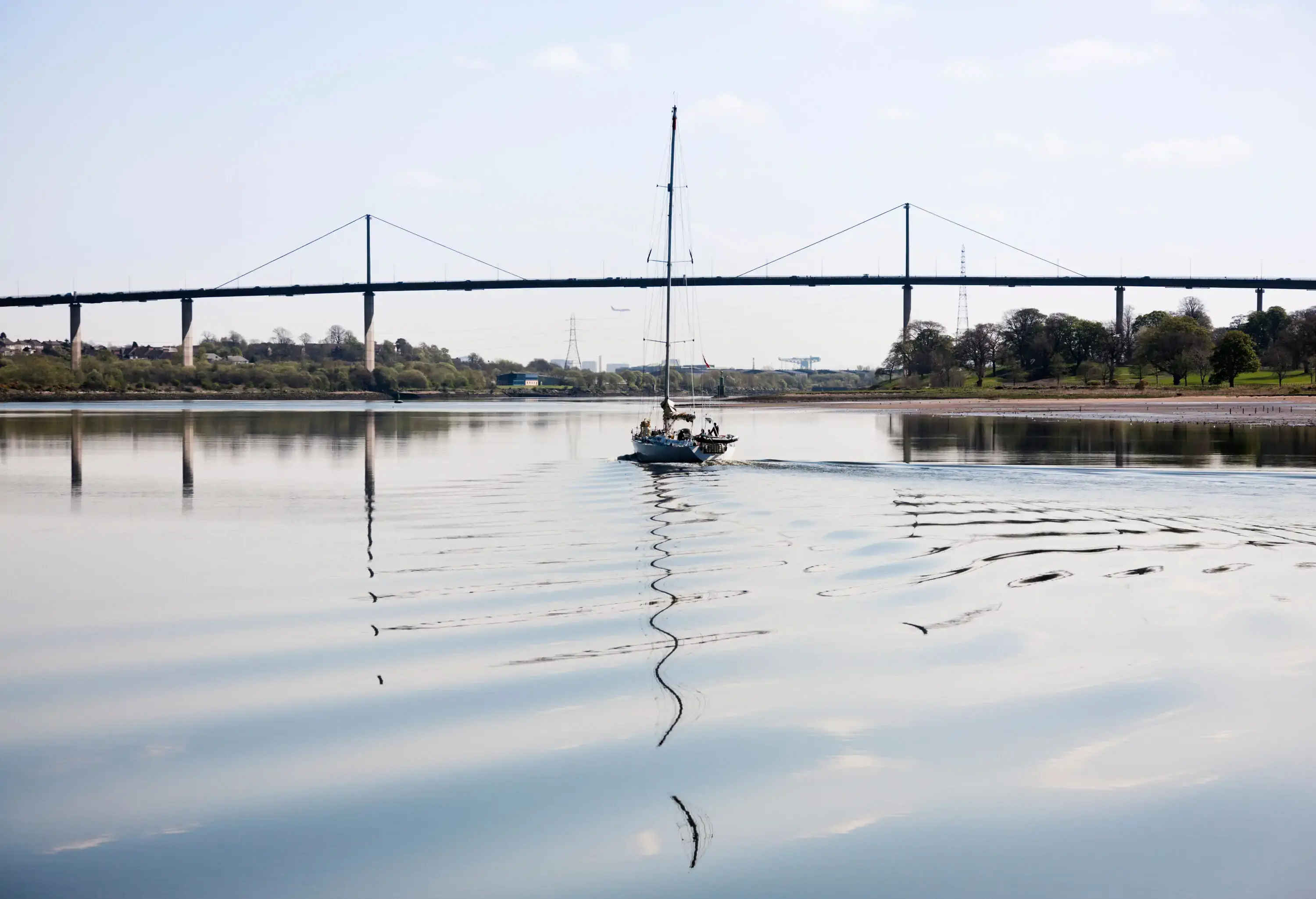 Sailing yacht sailing up the River Clyde with the Erskine Bridge behind and Glasgow beyond.