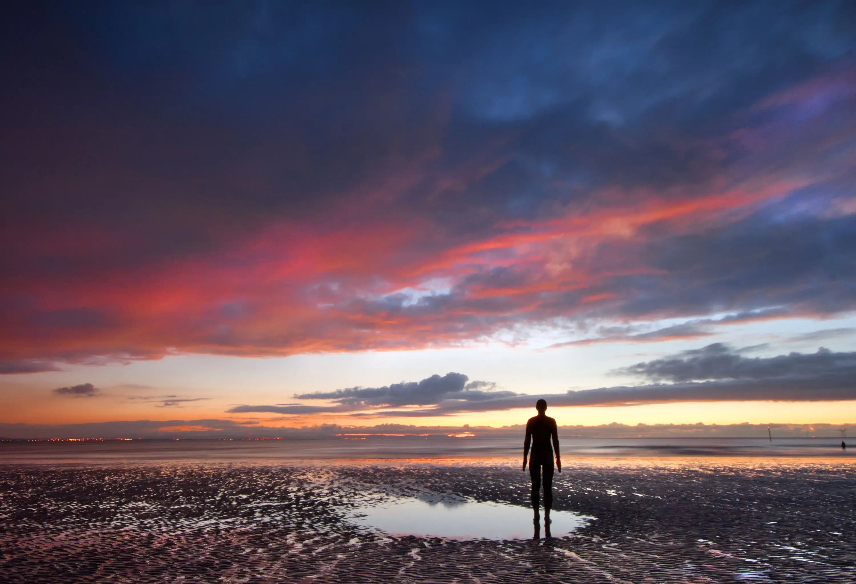 A lone figure gazing at the sea on a deserted beach by a pool of water at sunset
