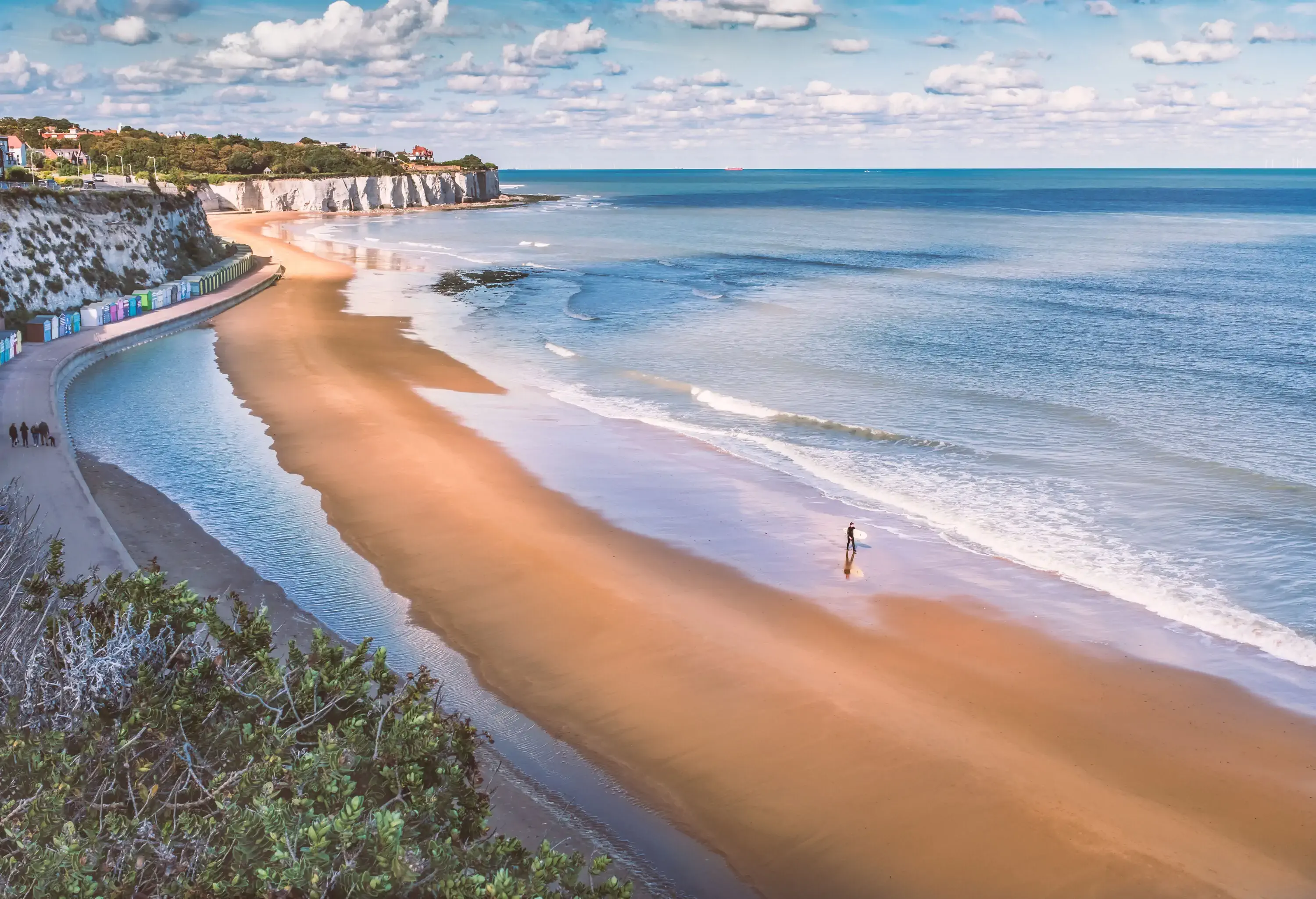 A lone surfer walks on a beach while holding his surfboard, with views of a distant white cliffs and a big ocean on the horizon.