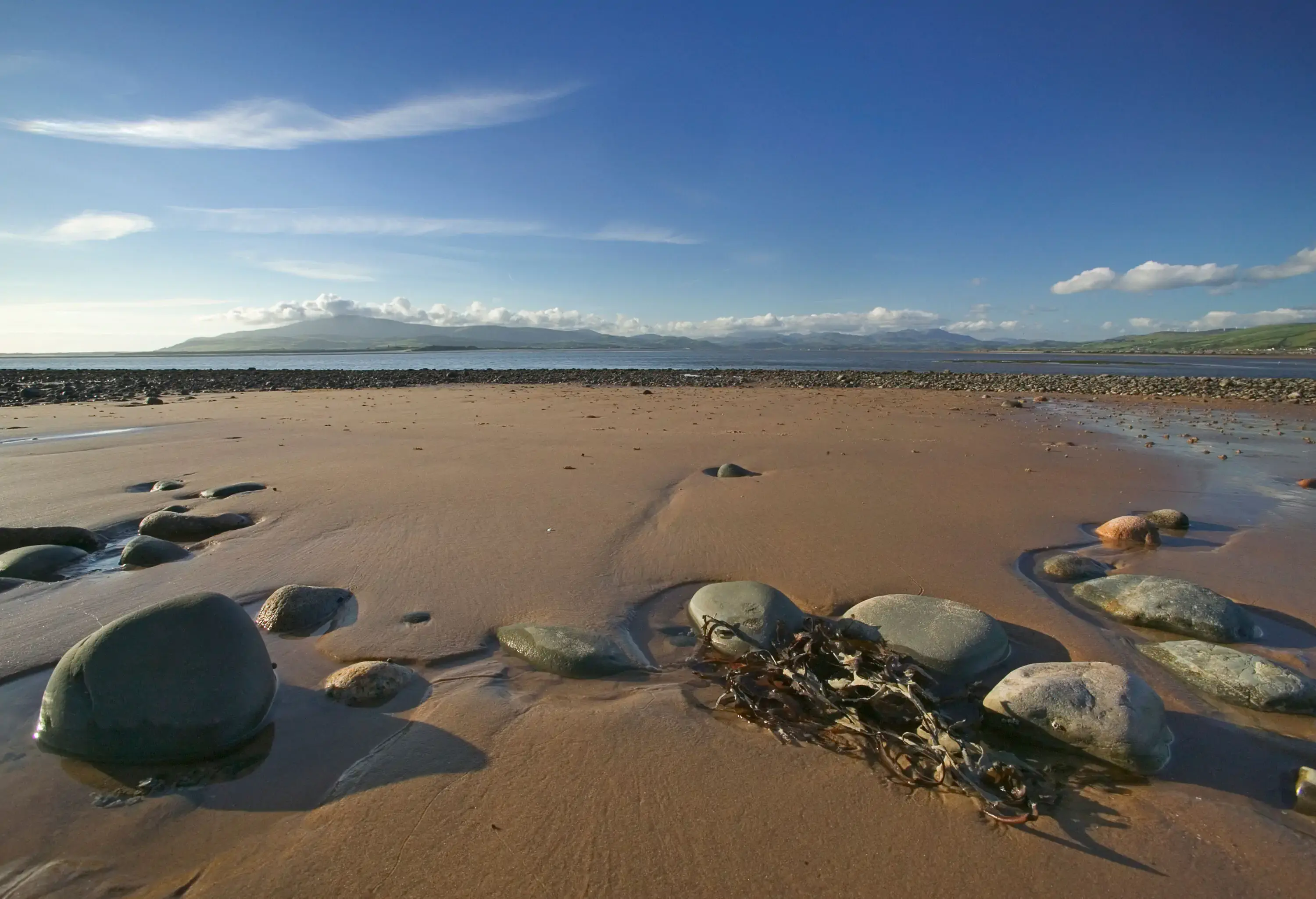 A wide sandy beach with stones in the foreground at low tide on a sunny day
