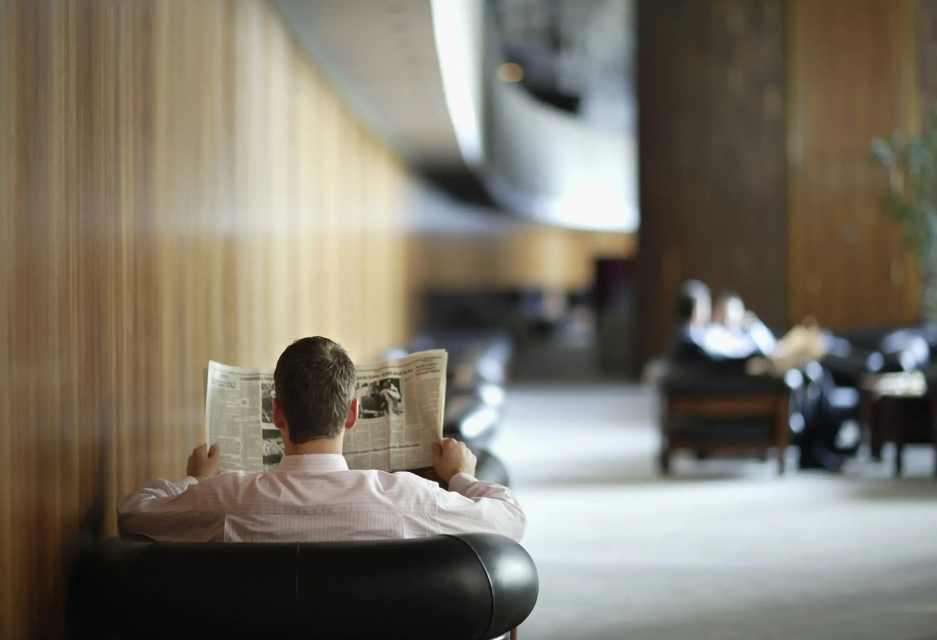 A man sitting on an armchair, reading a newspaper in a professional business environment with sleek decor.