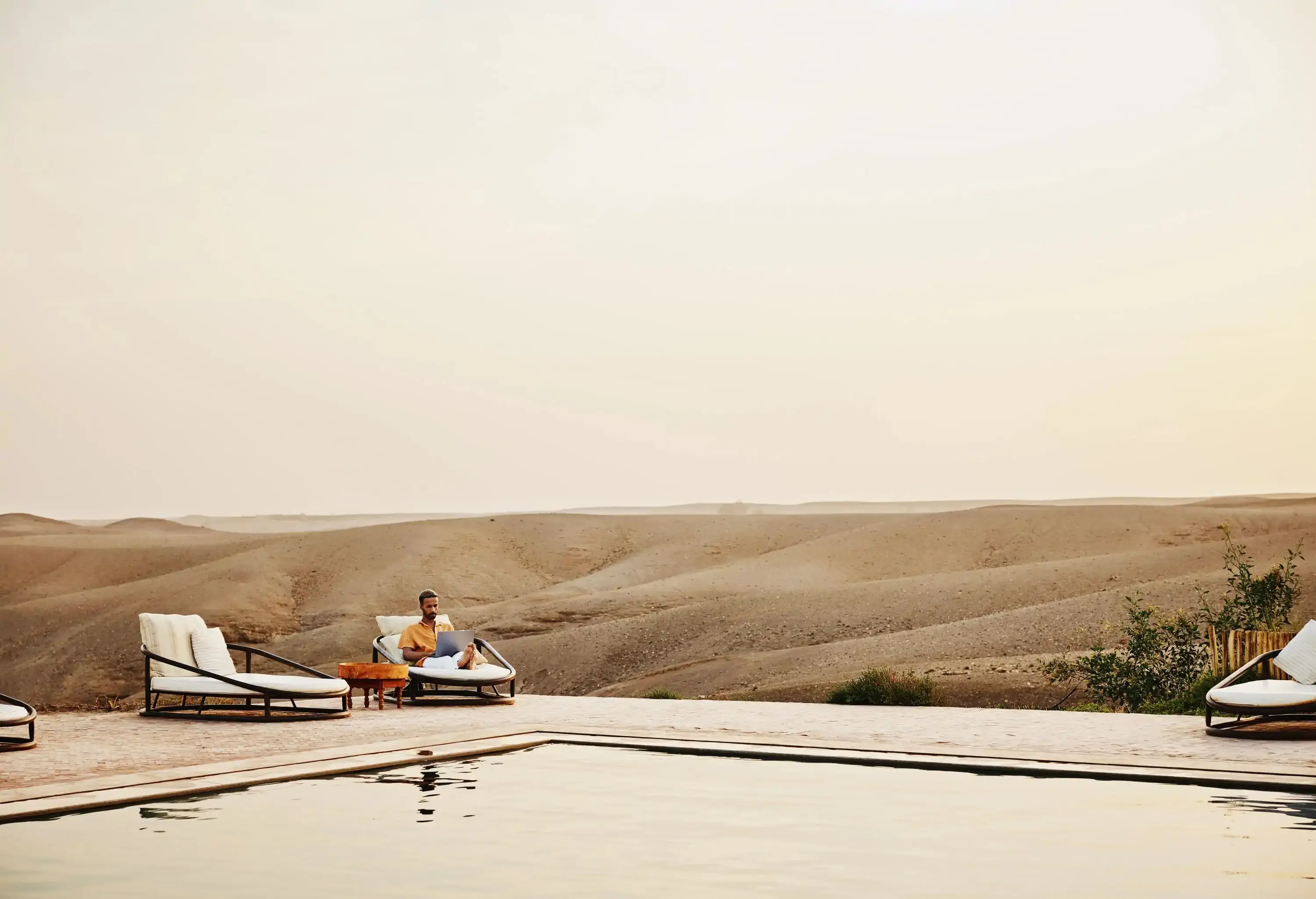 Wide shot of a man working on a laptop while sitting beside a pool at a Moroccan desert camp, surrounded by scenic desert landscapes during a vacation.