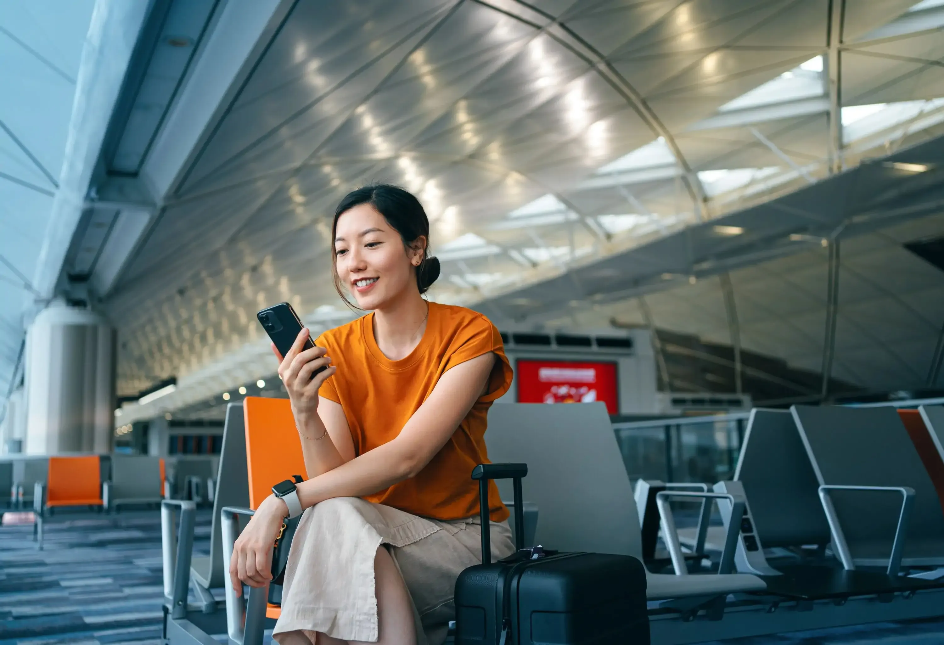 Young Asian woman with a suitcase, using a smartphone while seated in an airport terminal, waiting for her flight. Business traveler blending lifestyle and technology.