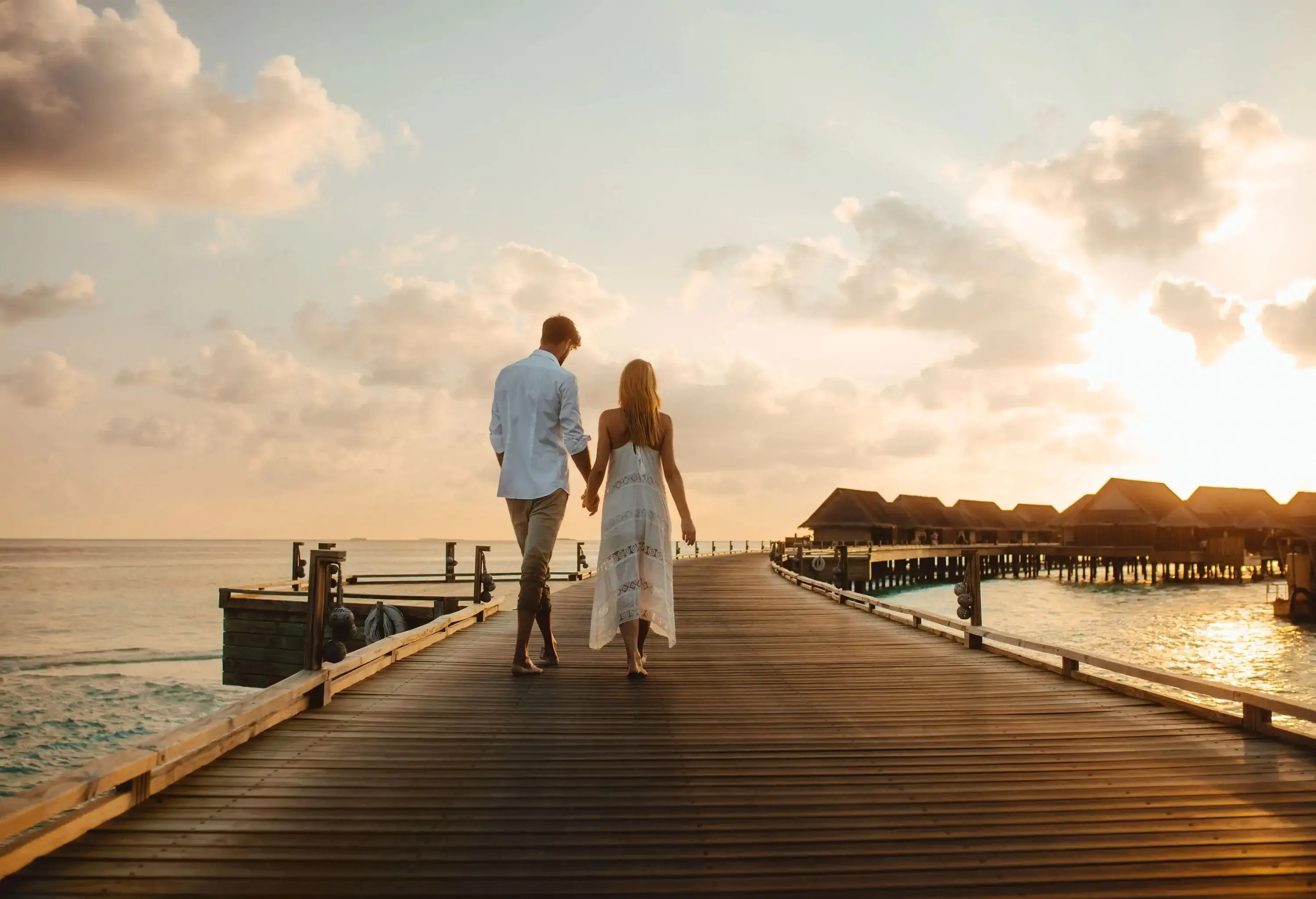 A tourist couple holds hands while walking on the boardwalk towards the floating resort villas on a sunset.