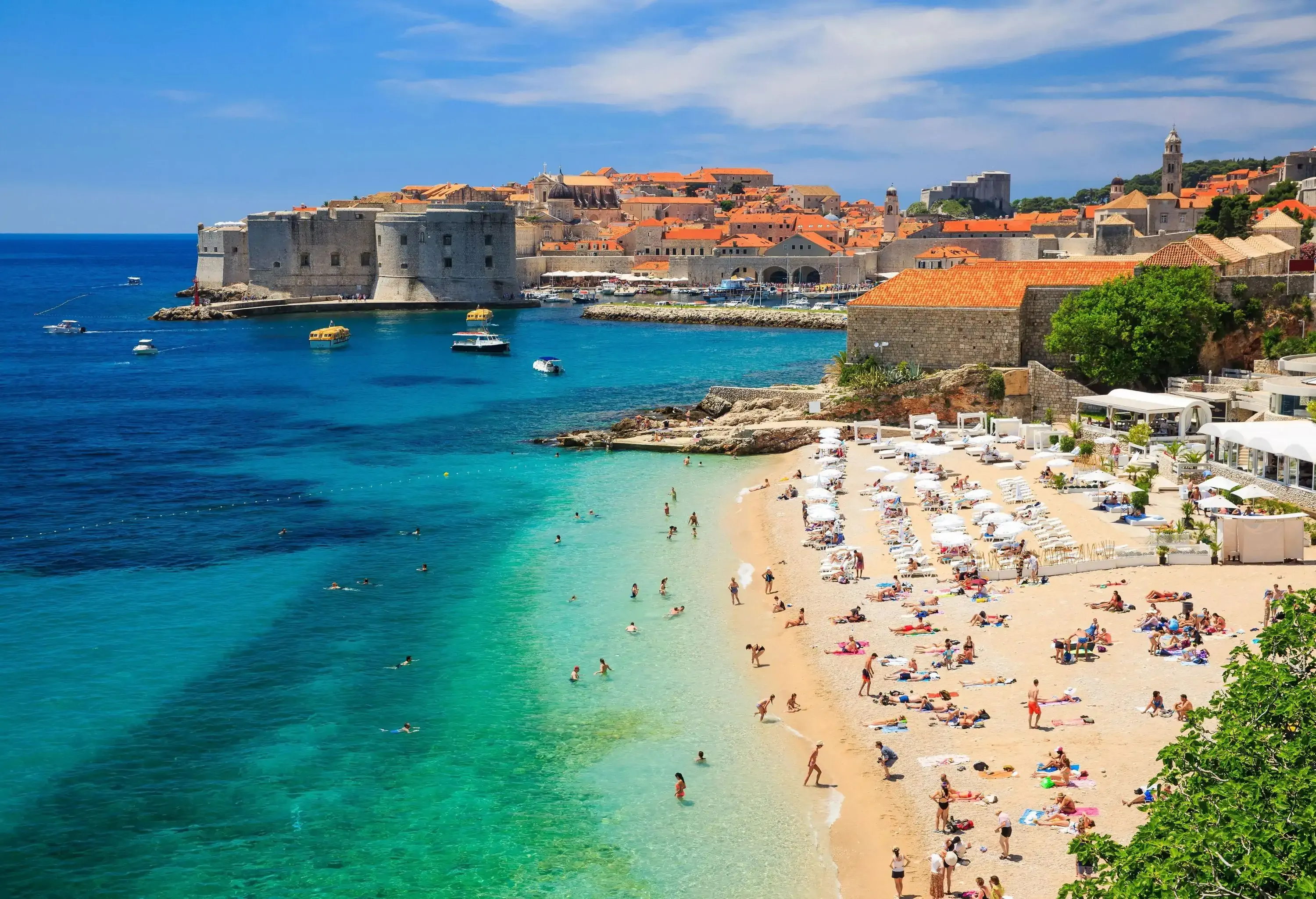 A busy beach with turquoise water and old fortified city in the background