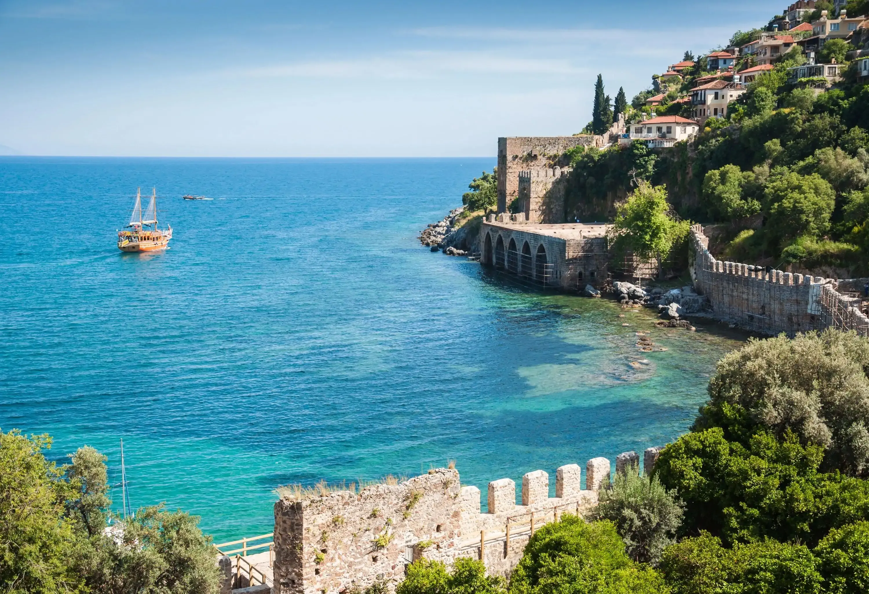 A medieval wall framing the coast, with buildings perched on a distant slope and a leisurely boat cruise nearby.
