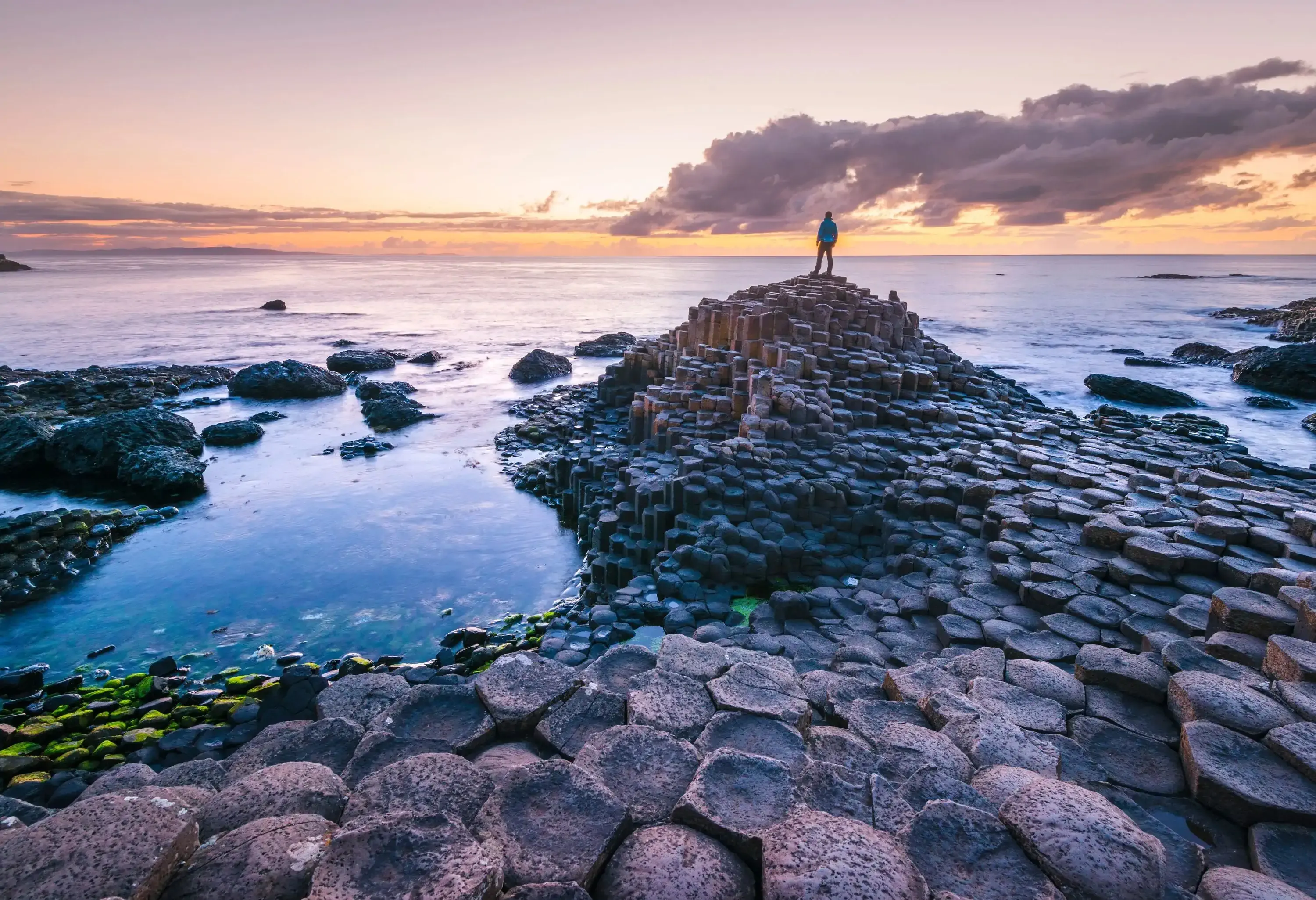 Person standing on rock formations looking at the ocean at sunset with purple overtones