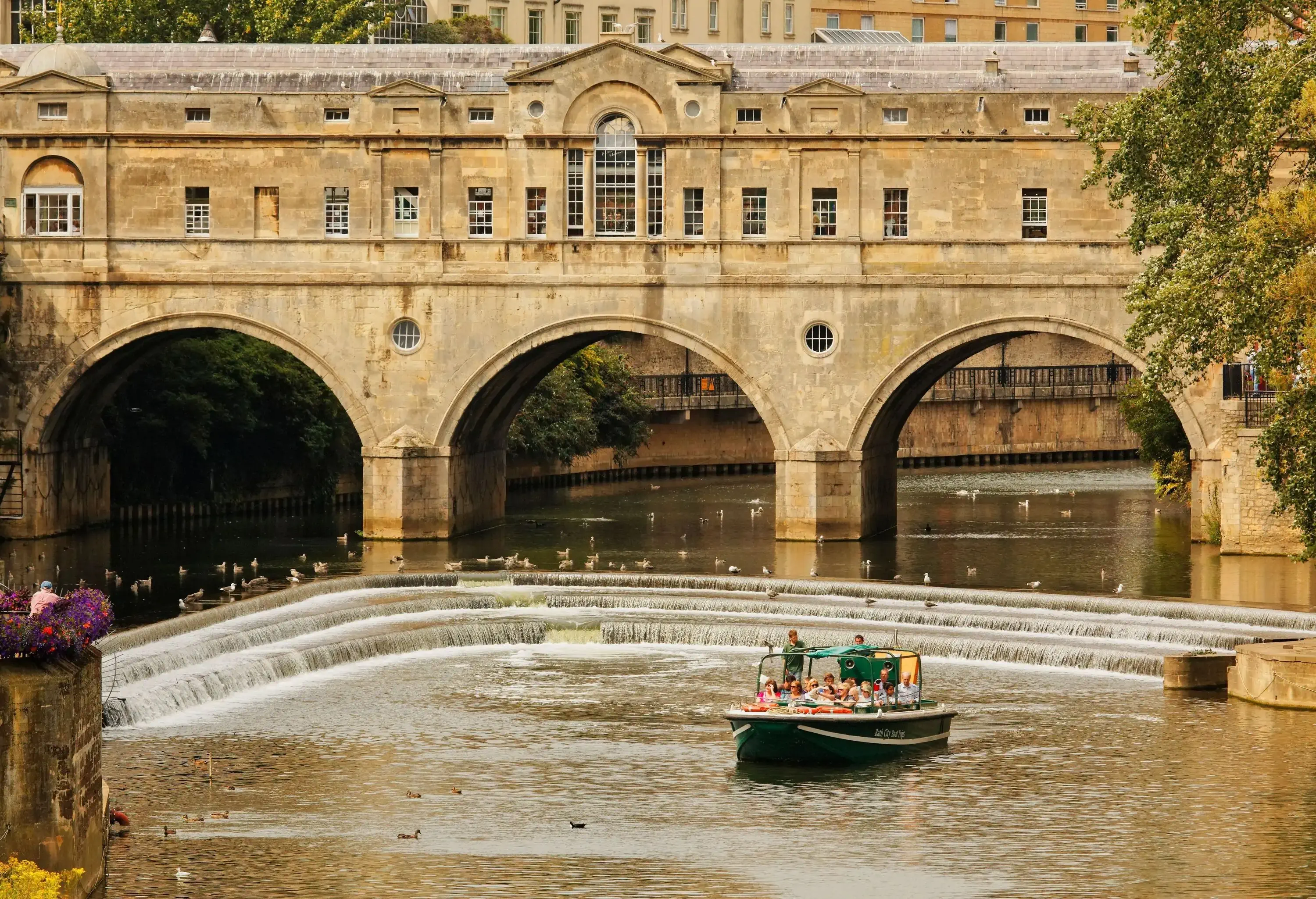A loaded boat crossing a weir in a river with an arch bridge in the background.