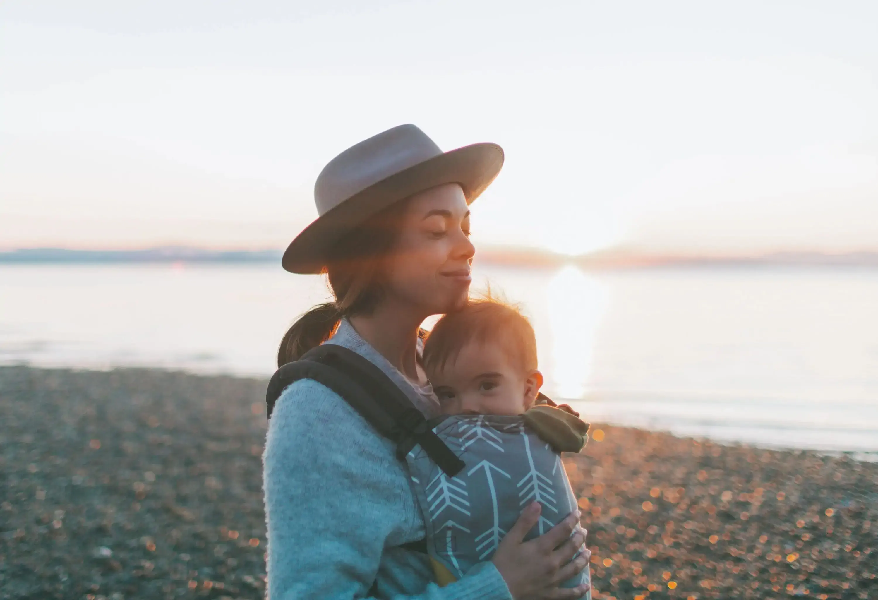 A mother in a hat hugs her baby while standing on a beach.