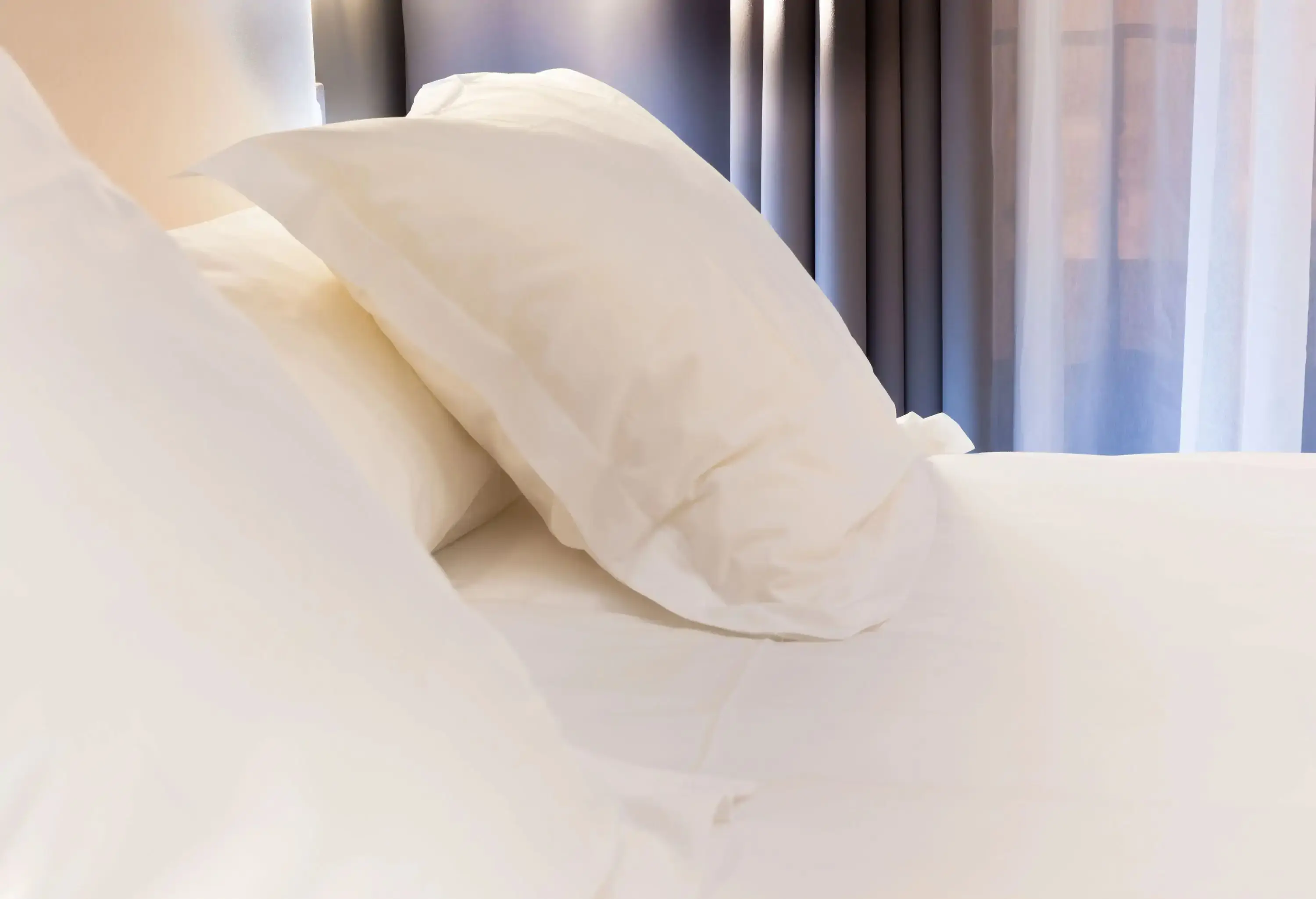 Clean white bedsheets and pillows inside a cosy bedroom with greyish curtains.