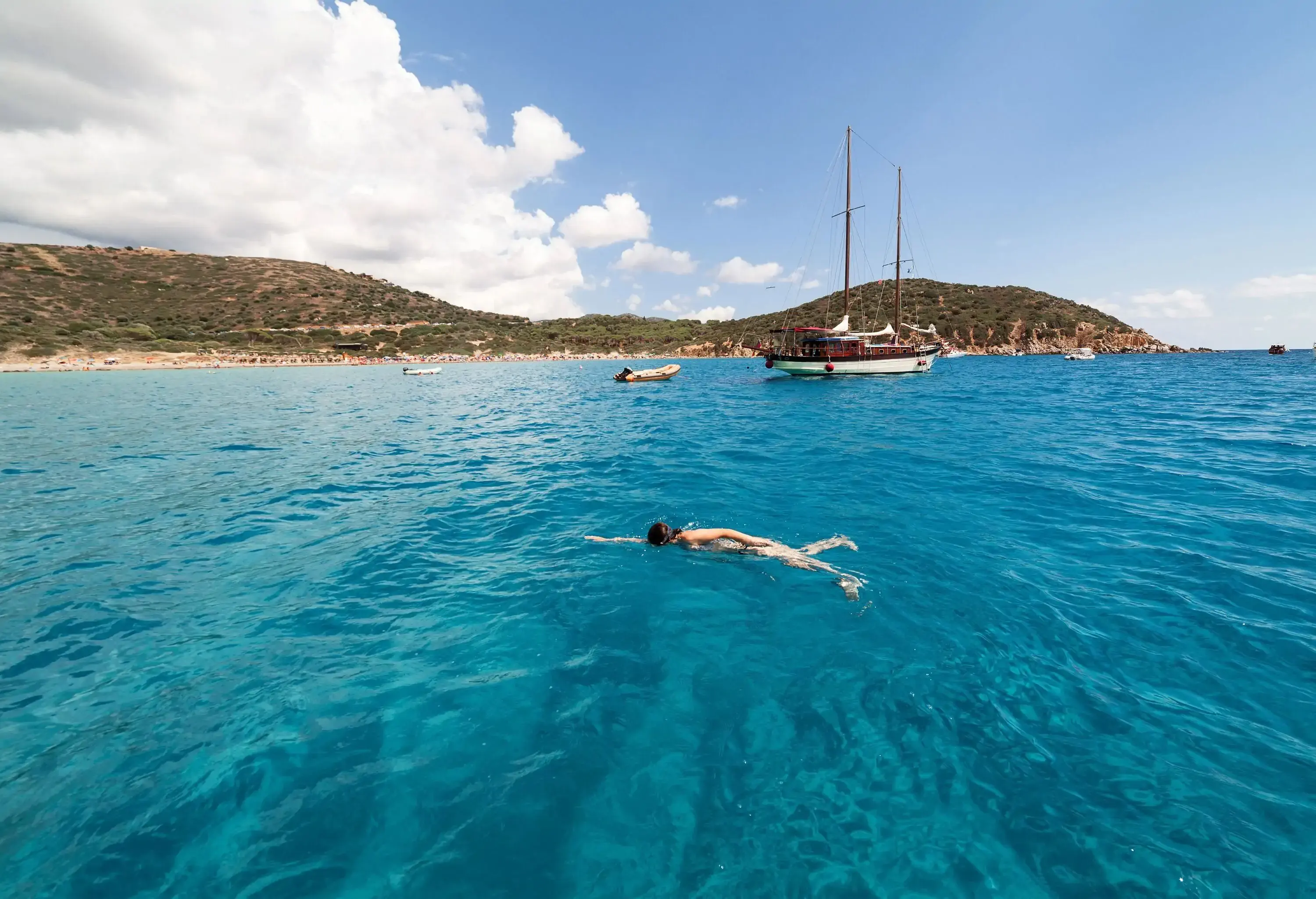 A captivating seascape features a woman gracefully swimming in the turquoise sea, accompanied by boats, while verdant slopes adorn the shore.