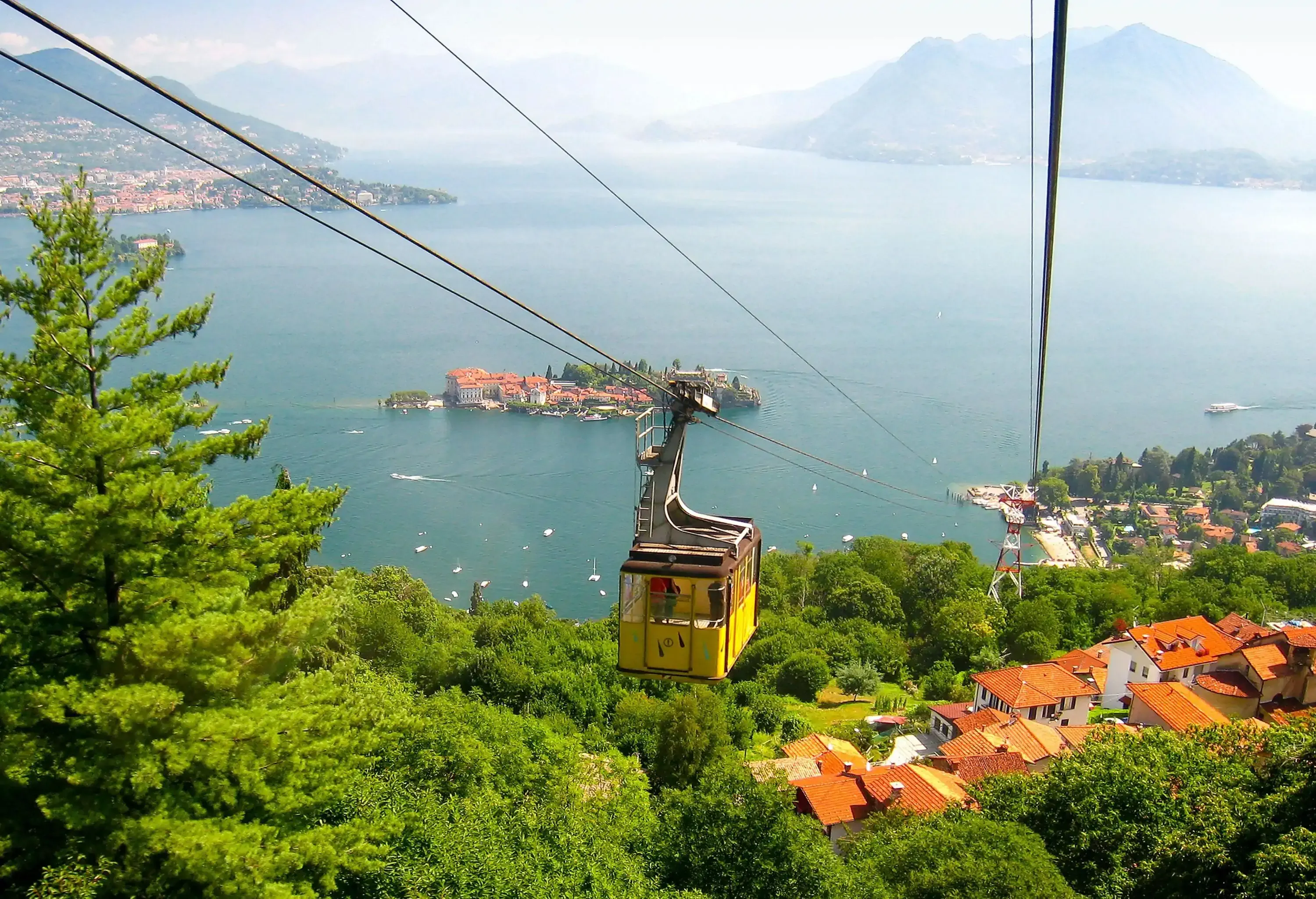 A yellow cable car travelling over village houses and trees by a lake.