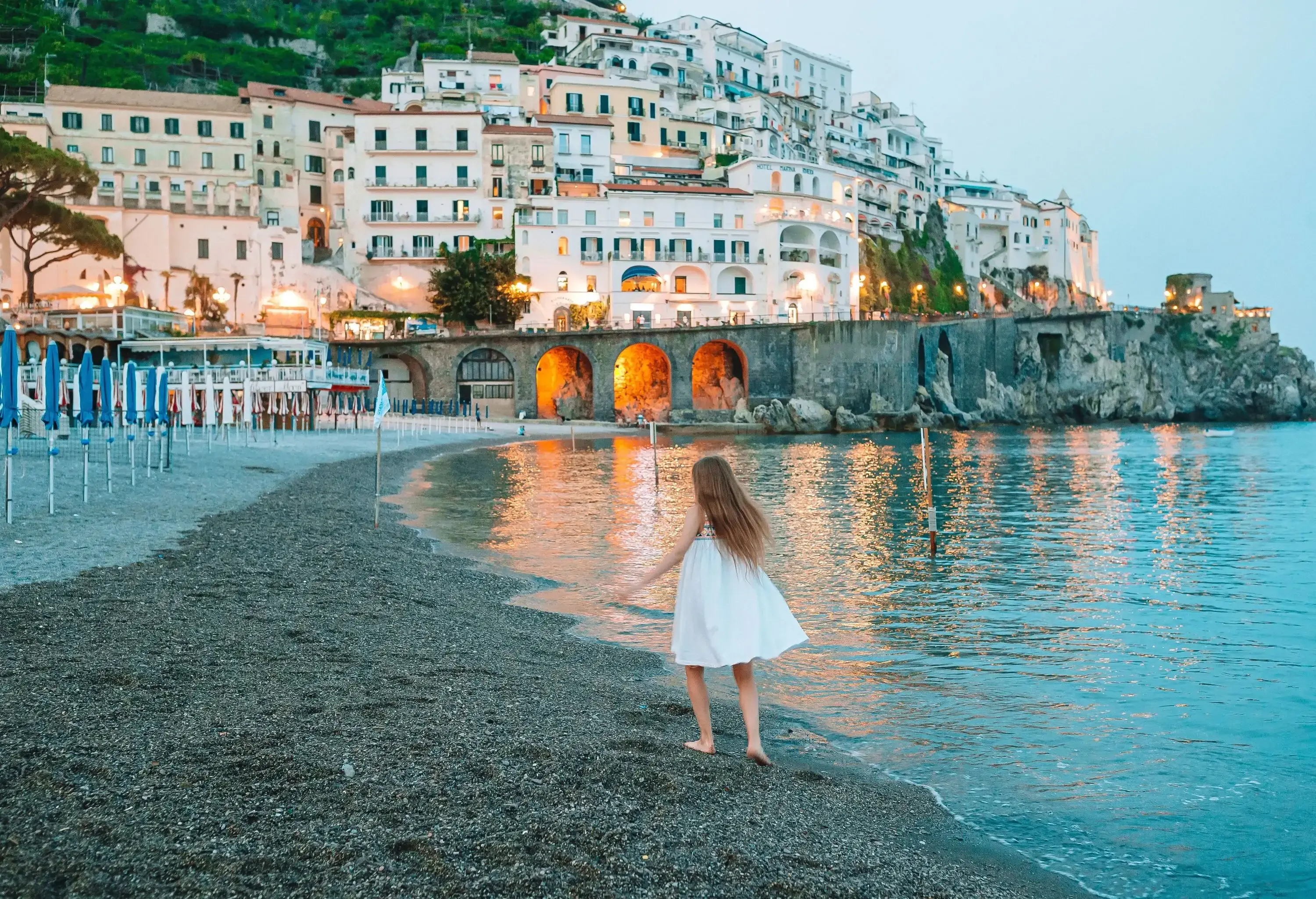 Girl in a white dress strolls down a beach, with distant views of white buildings situated on the cliff's edge.
