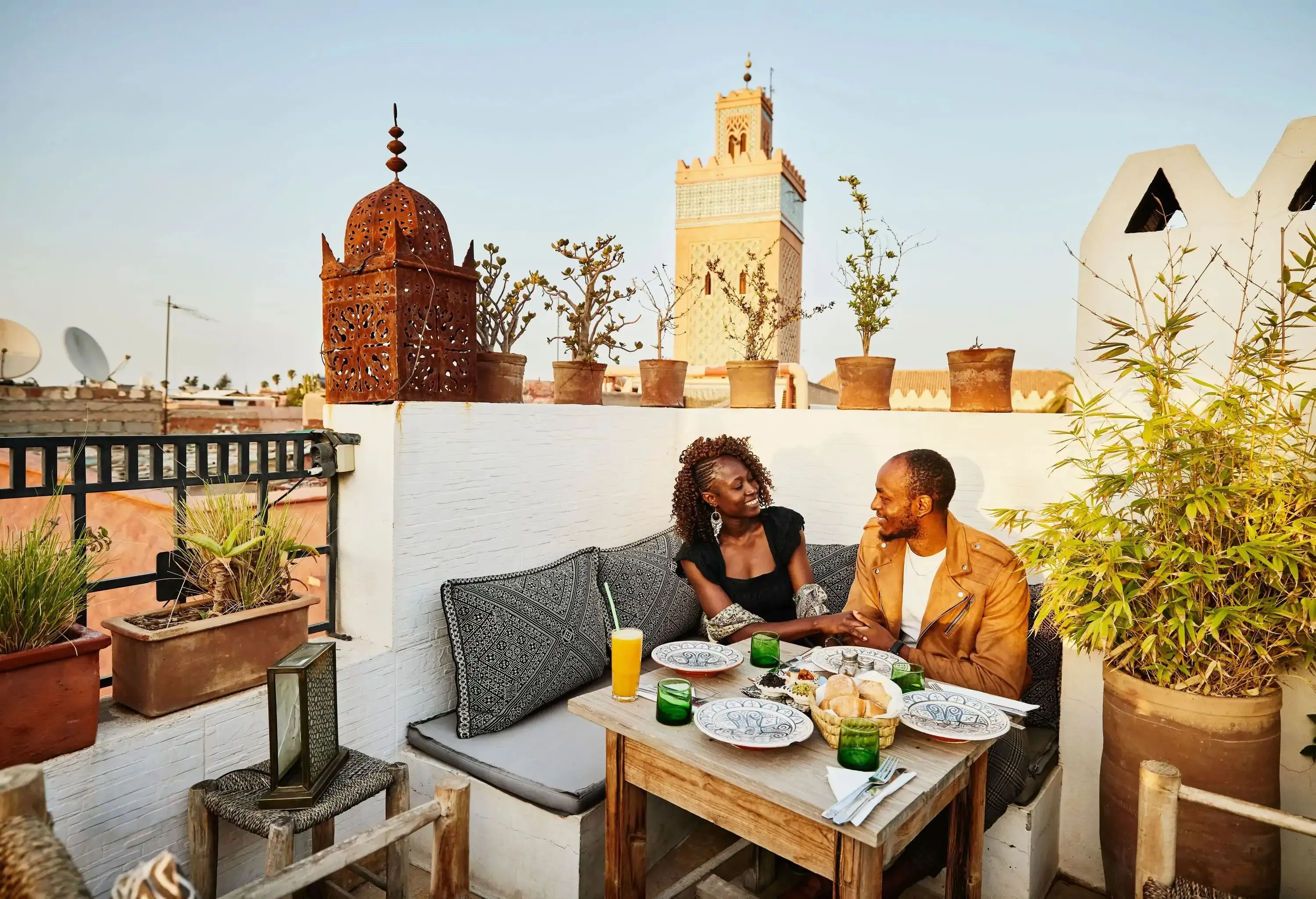Couple enjoying a meal on a rooftop terrace surrounded by potted plants, with a tall minaret and decorative structures in the background.