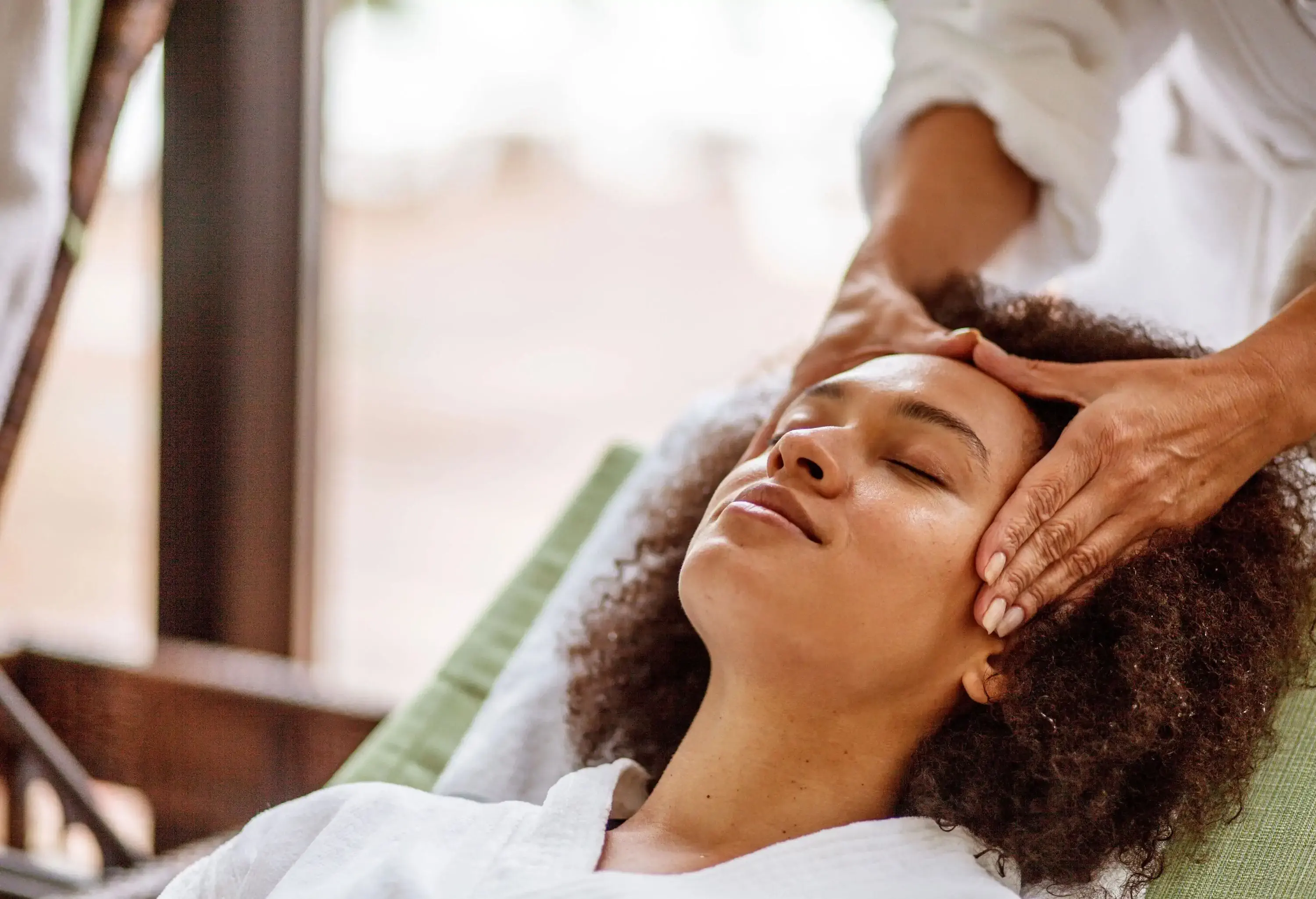A curly-haired woman receives a head massage with her eyes closed.