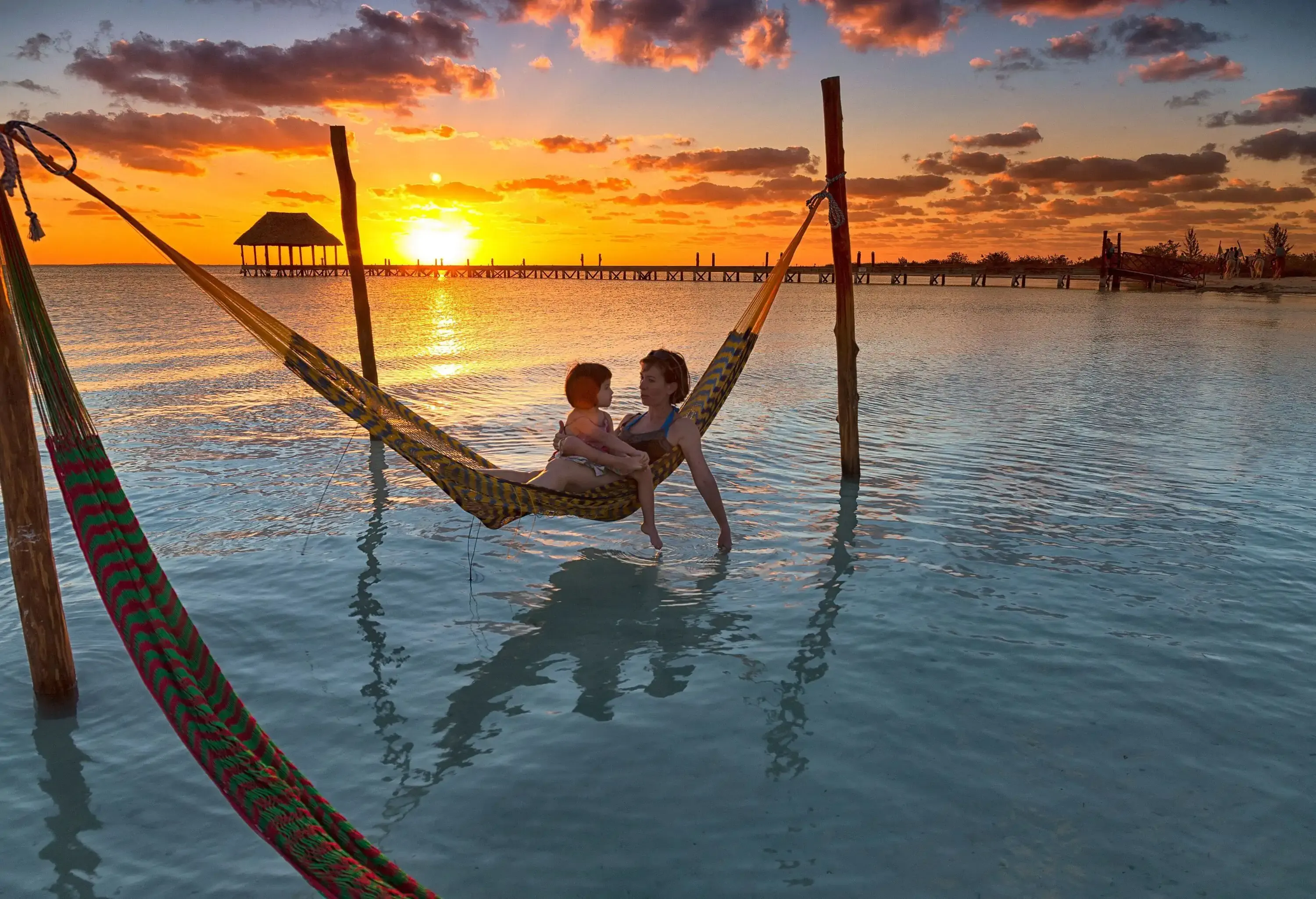 A scenic view of a mother and daughter on a hammock at the beach against the dramatic sunset sky.