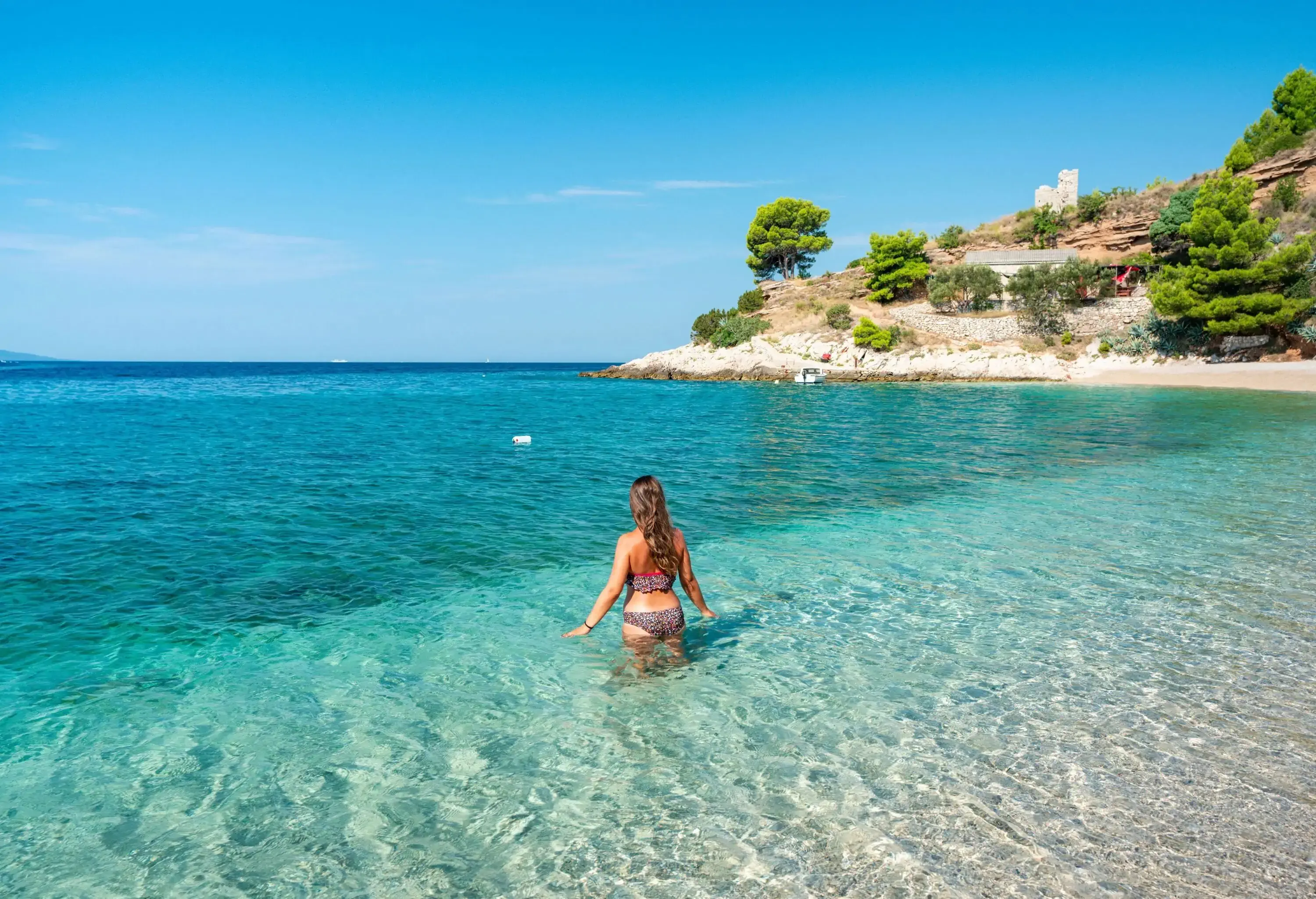 Half of a woman in a bikini is submerged in clear water near a beach with rocky shores. 