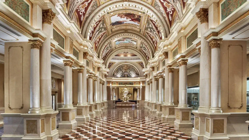 Ornate, high-ceiling corridor with marble columns, intricate frescoes, and a reflective checkered floor. A grand, opulent atmosphere is conveyed.